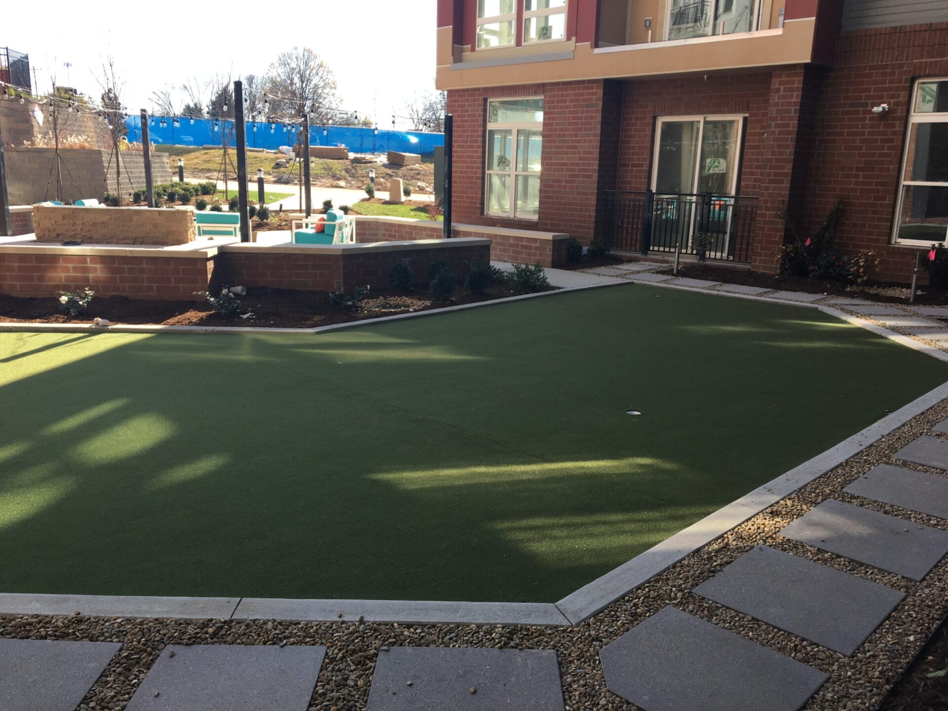 This image shows a small outdoor putting green bordered by brick, with a path of stepping stones leading up to it, near a red brick building.
