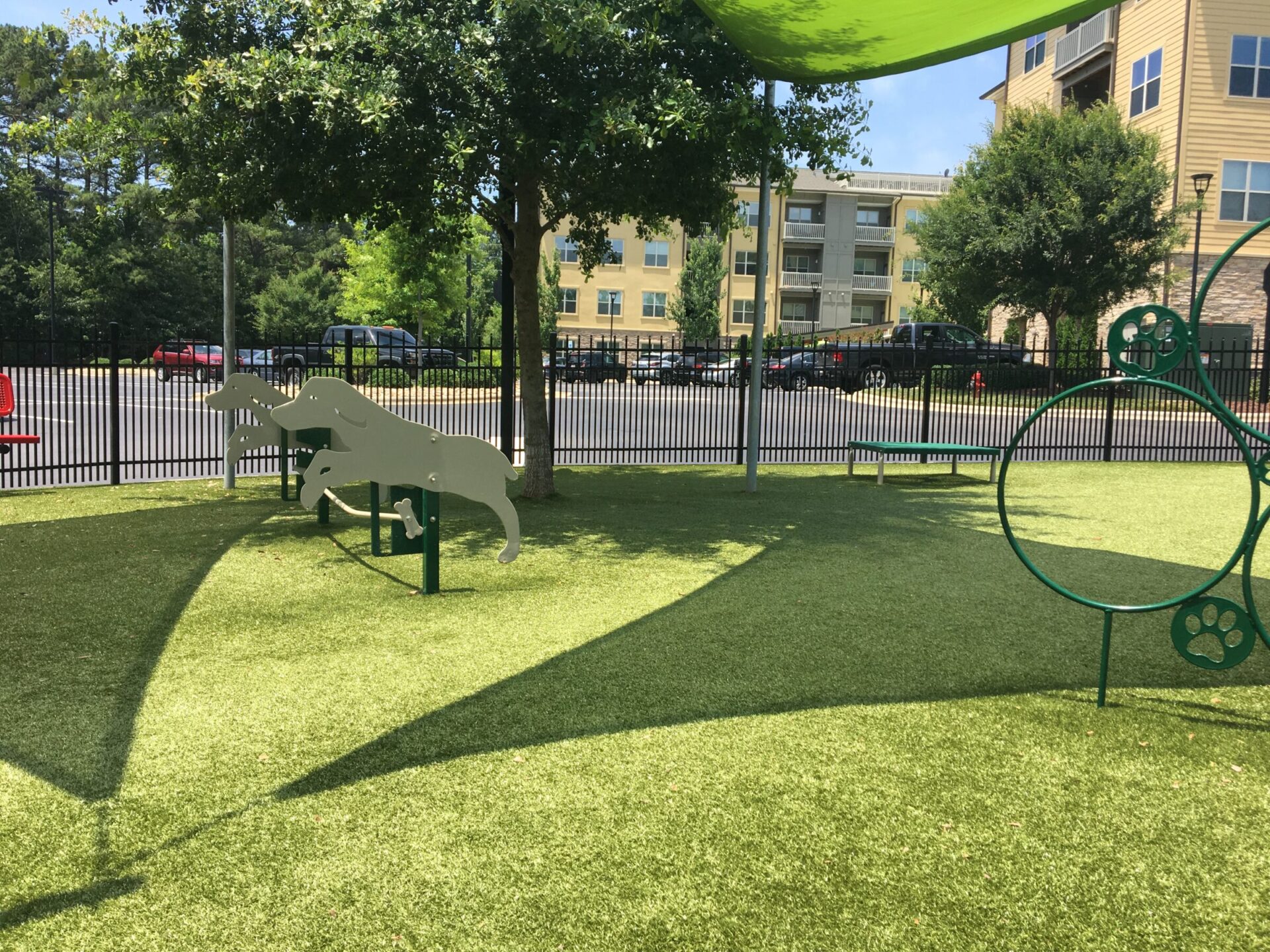 A sunny playground with artificial grass features a green dinosaur structure, agility rings, and a shaded area, surrounded by a fence with buildings behind.