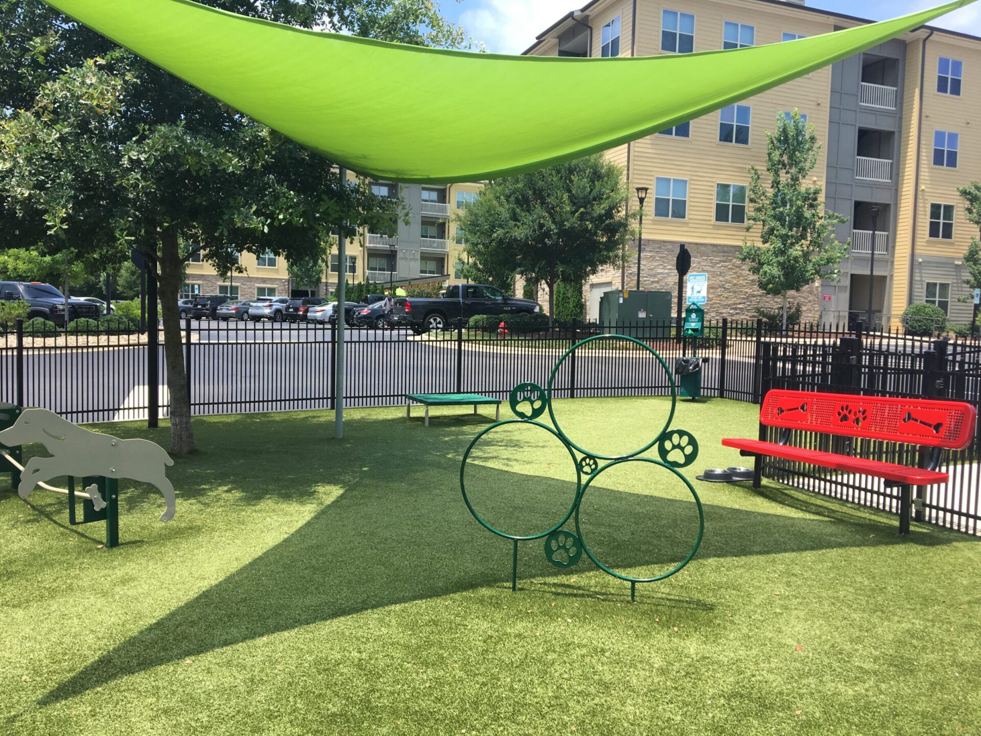 A sunny dog park with artificial grass, featuring agility equipment, benches with canine motifs, and a green shade sail. Apartment buildings are visible in the background.