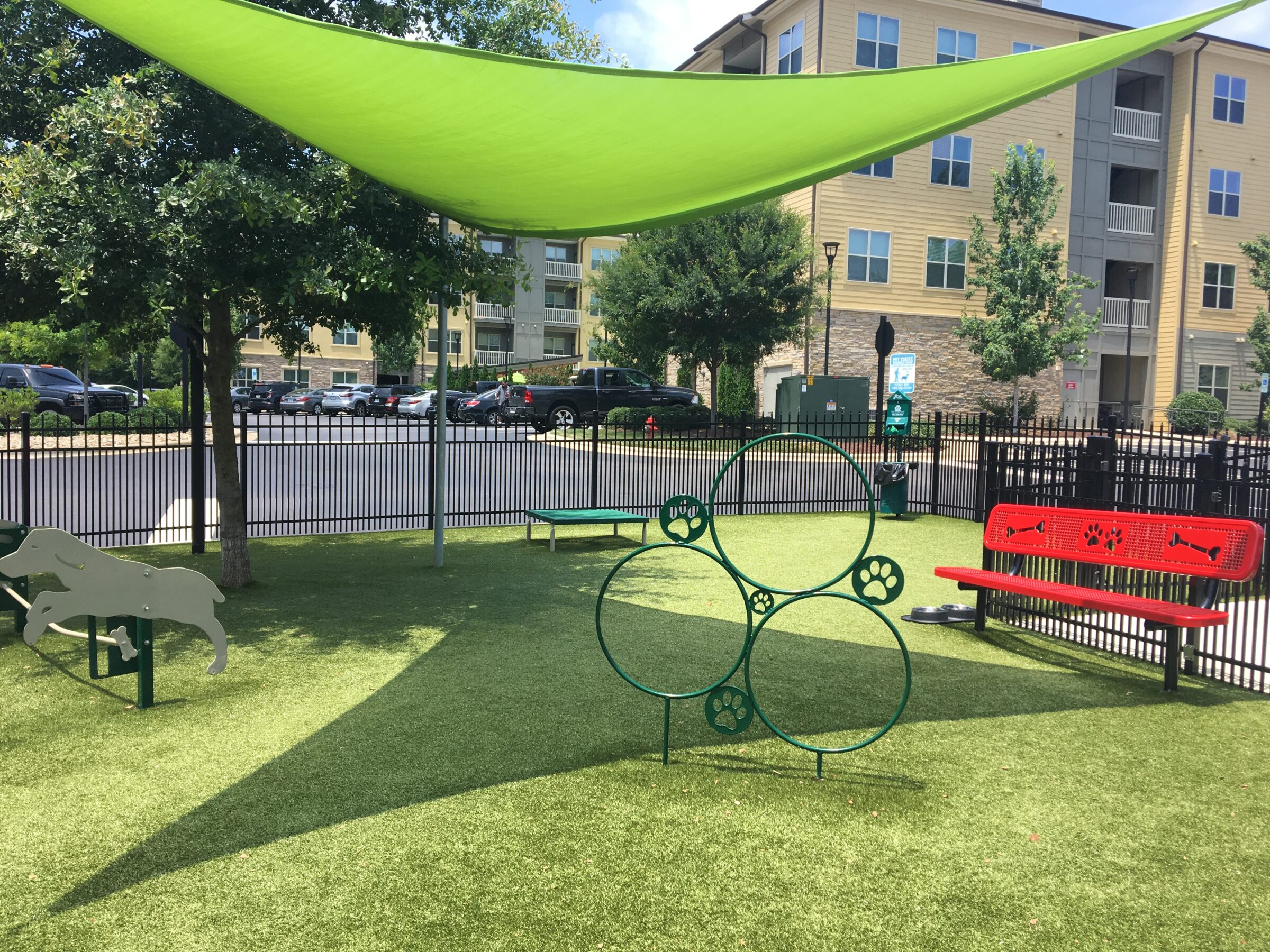 A sunny dog park with green synthetic grass, agility equipment, shaded benches, a water station, and a surrounding fence with apartment buildings in the background.