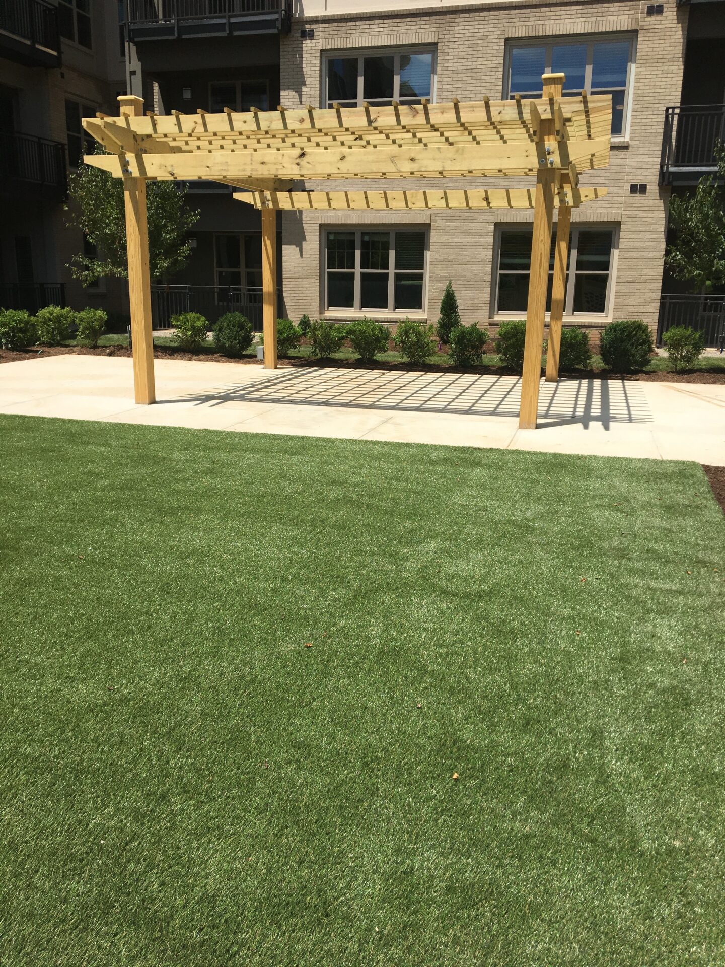 The image shows a well-manicured green lawn in front of a residential building with a modern pergola and a patterned pavement area underneath.