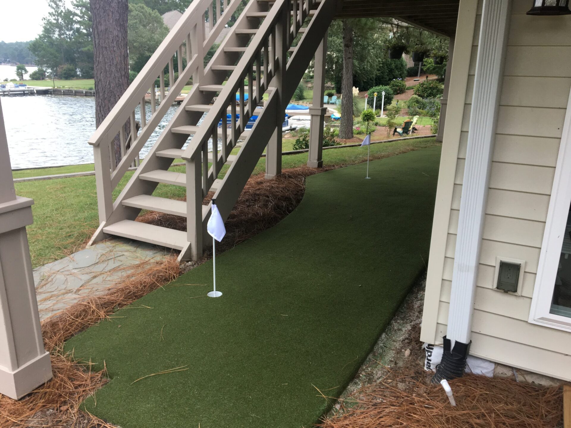 A backyard putting green alongside a house with stairs leading to a dock, flagsticks in holes, pine straw borders, and a lake view.