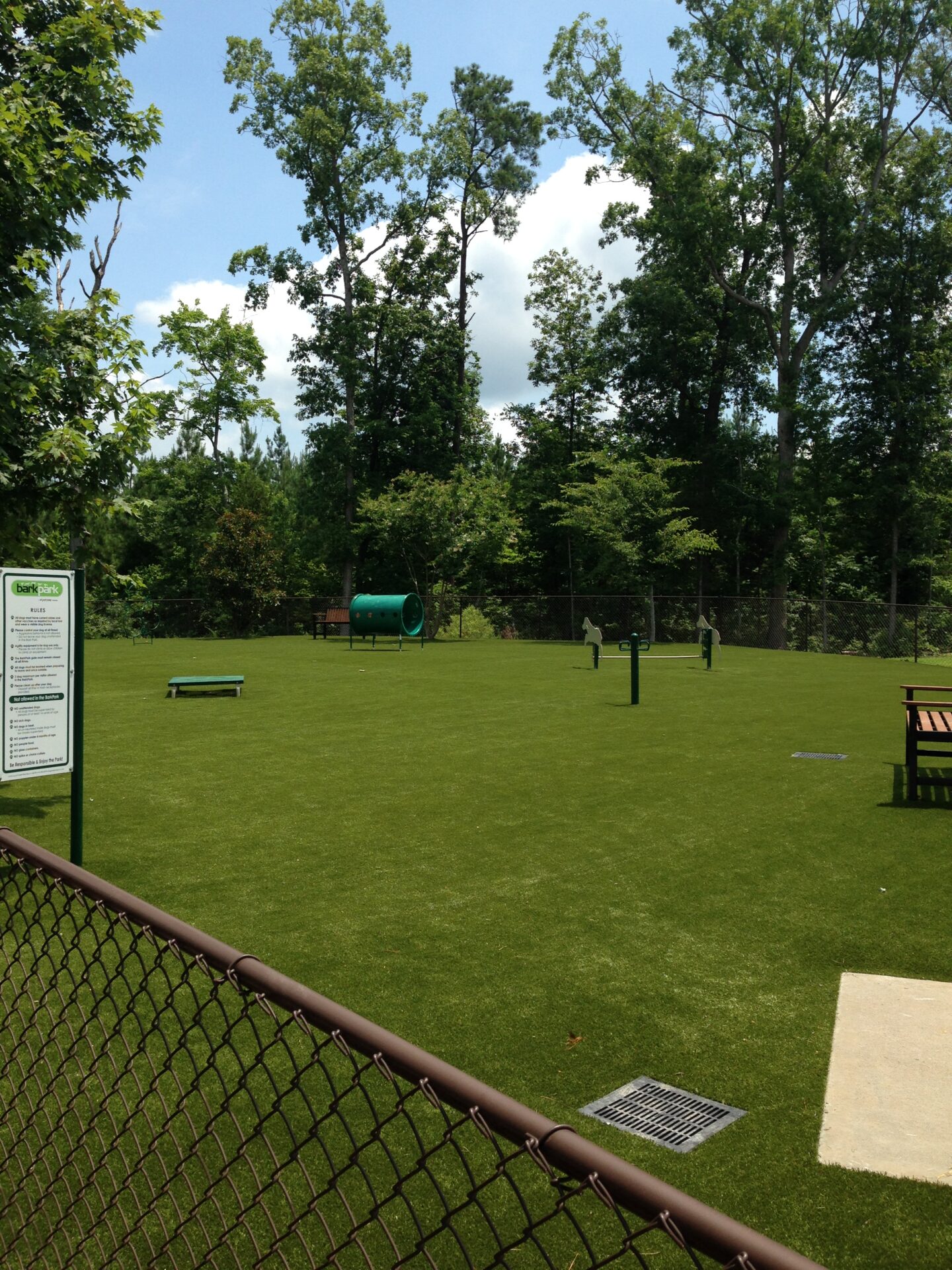 A dog park with artificial grass, surrounded by a chain-link fence, trees in the background, benches, and agility equipment on a sunny day.