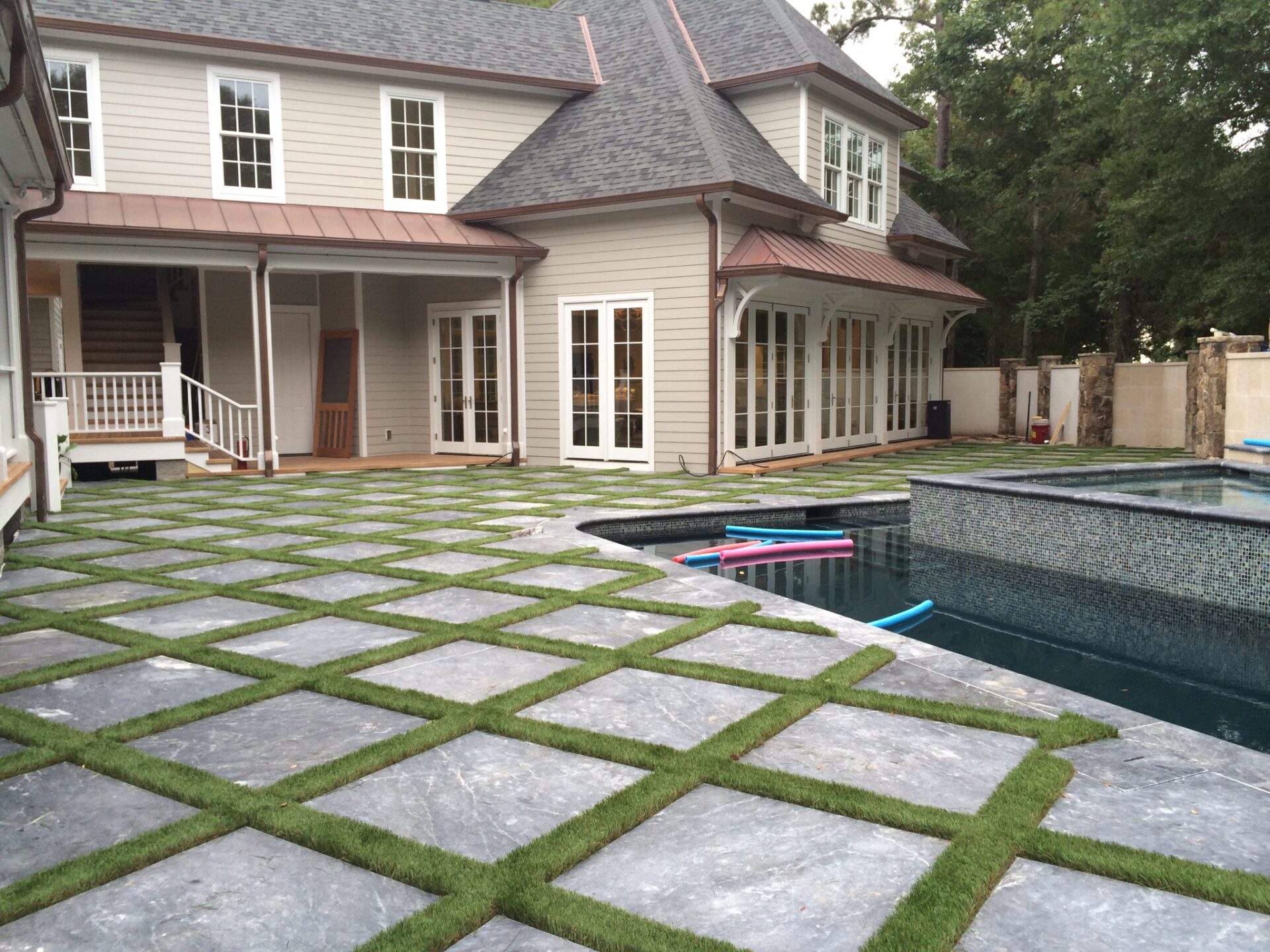 A large house with a slate tile patio and pool. The pool has floating foam noodles. There's grass between the patio tiles, and trees in the background.