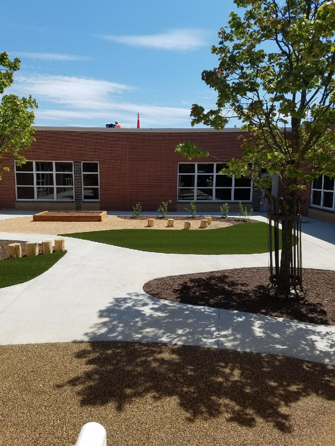 The image shows an outdoor courtyard with a brick building, green lawn areas, concrete walkways, wooden benches, and a growing tree. It's a sunny day.