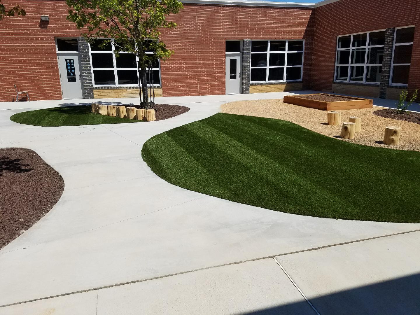 This image shows a landscaped outdoor area with synthetic grass, concrete walkways, wooden stumps for seating, a raised planter box, and a young tree.