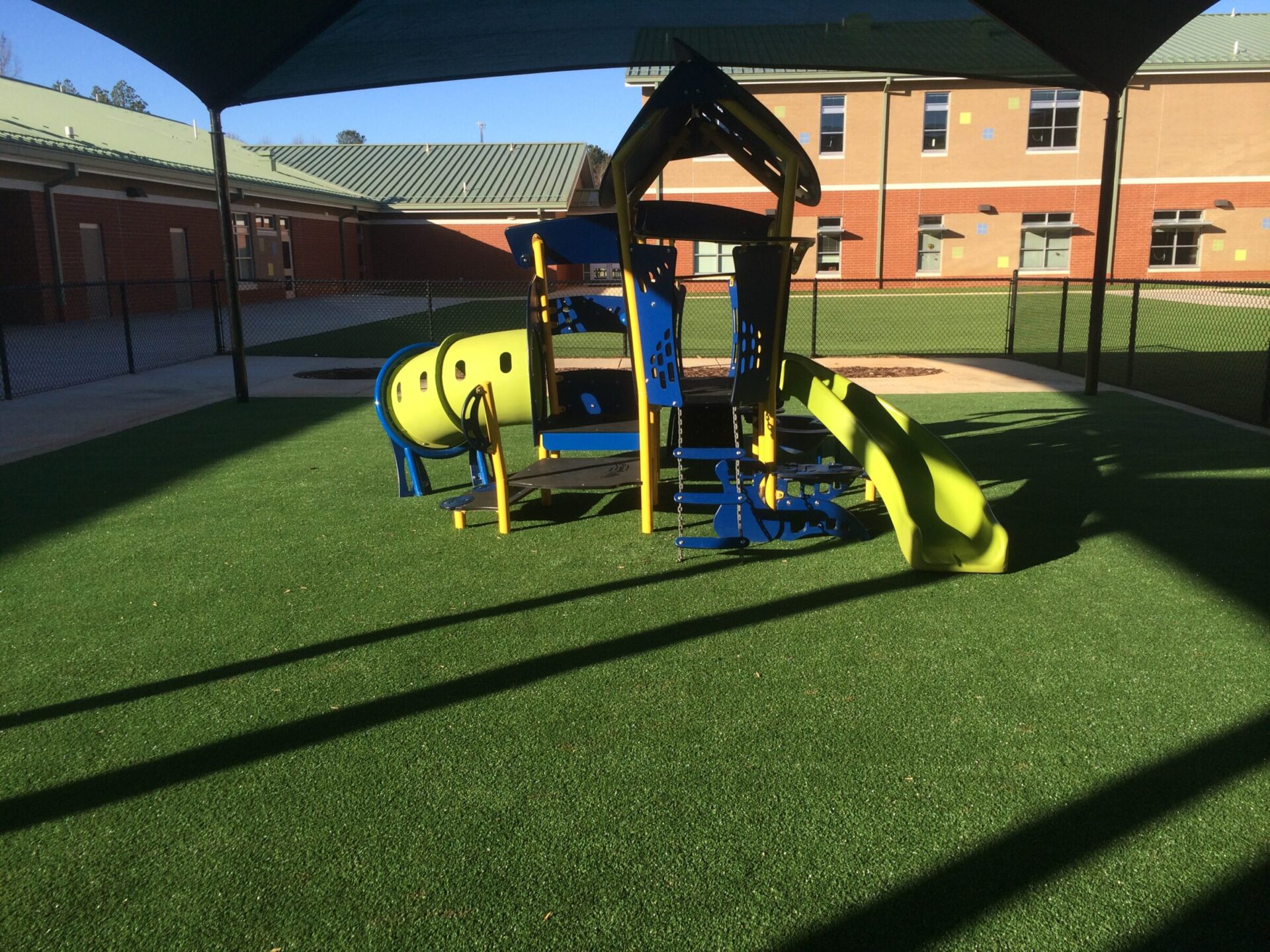 This image shows a colorful playground with a slide and climbing structures on artificial grass, shaded by a canopy, with school buildings in the background.