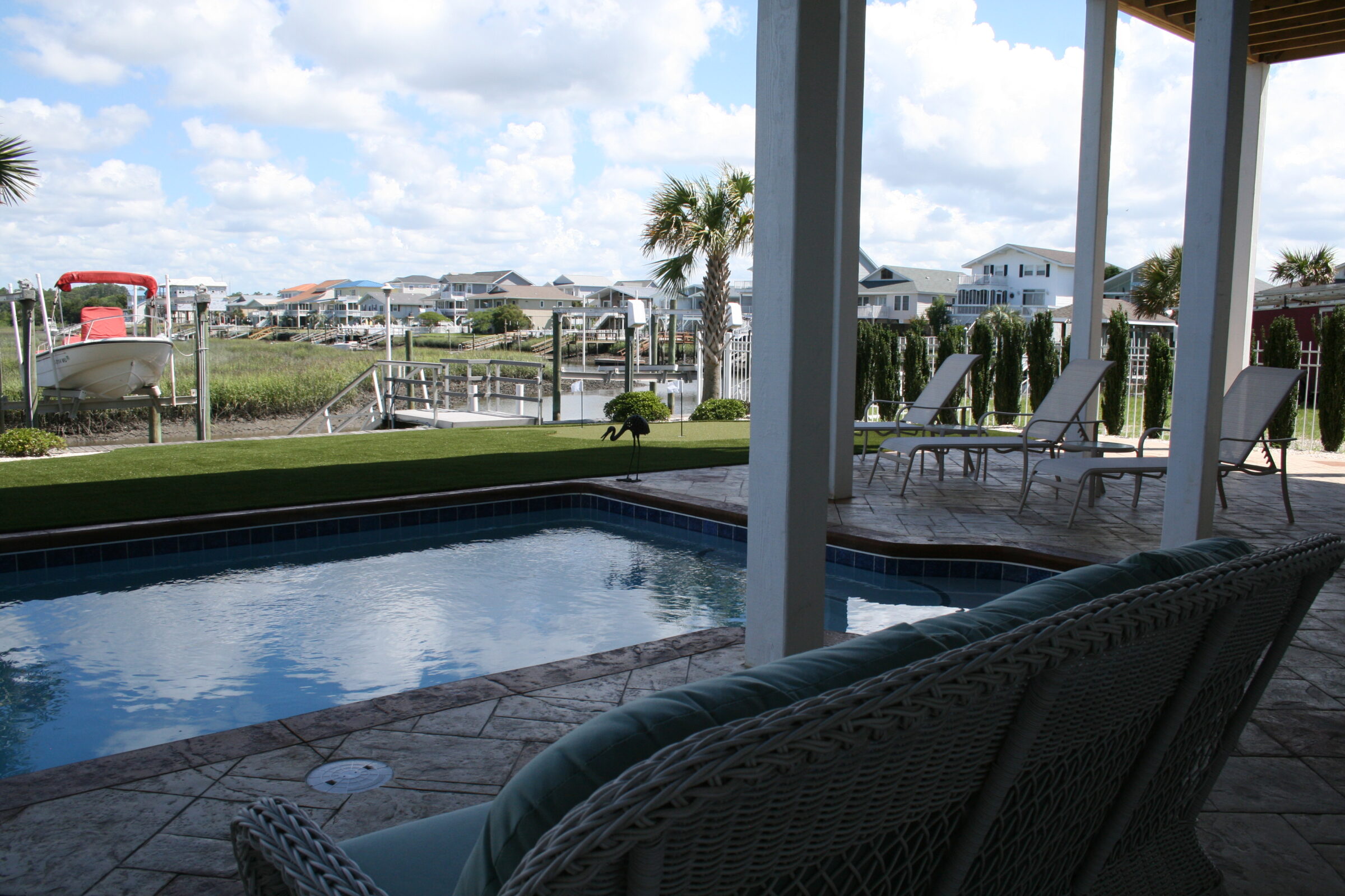 A serene backyard with a swimming pool, patio seating, and a boat docked by the waterway under a clear sky in a residential area.