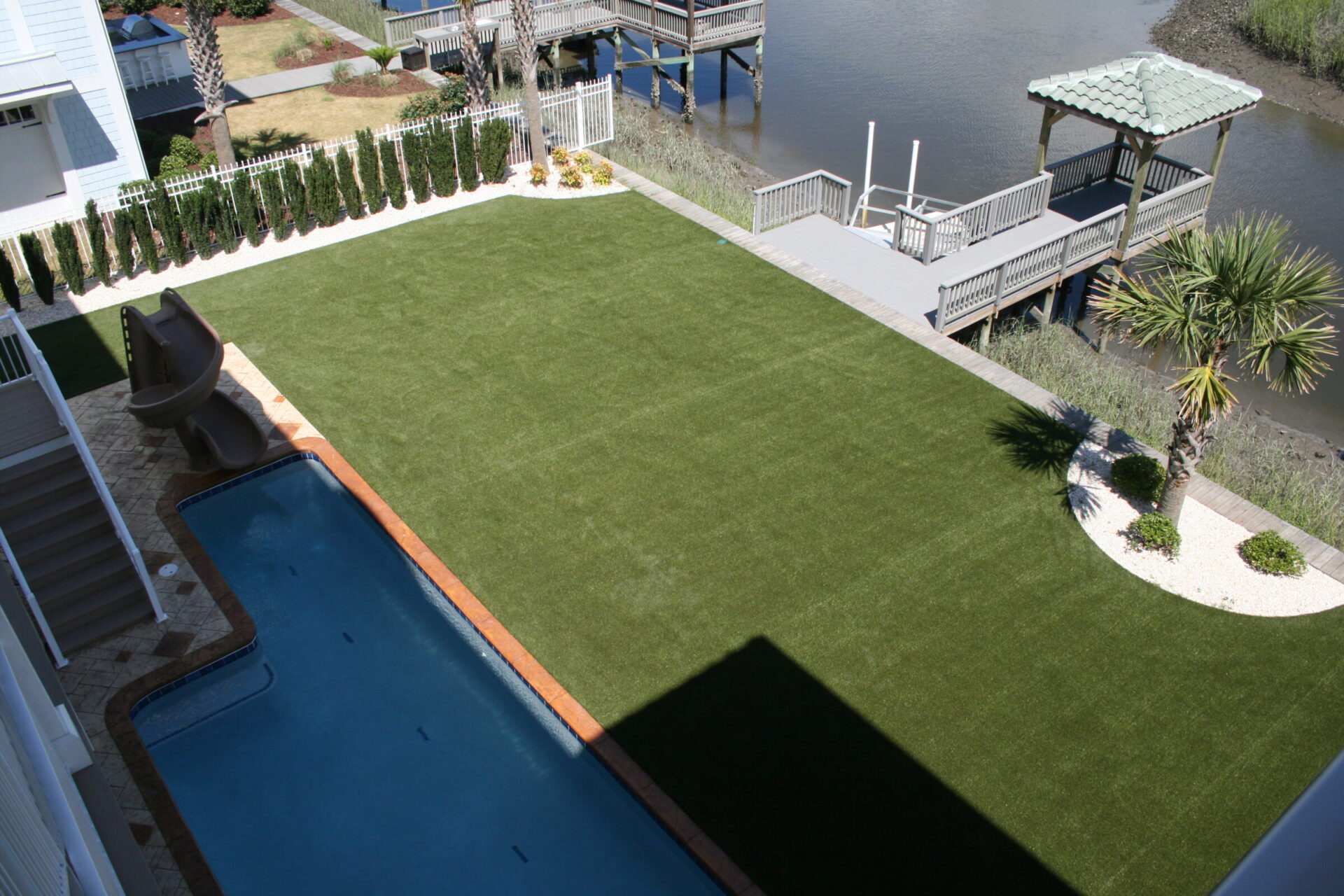An aerial view of a backyard with a swimming pool, slide, artificial grass, a palm tree, a gazebo, dock over water, and a fence.