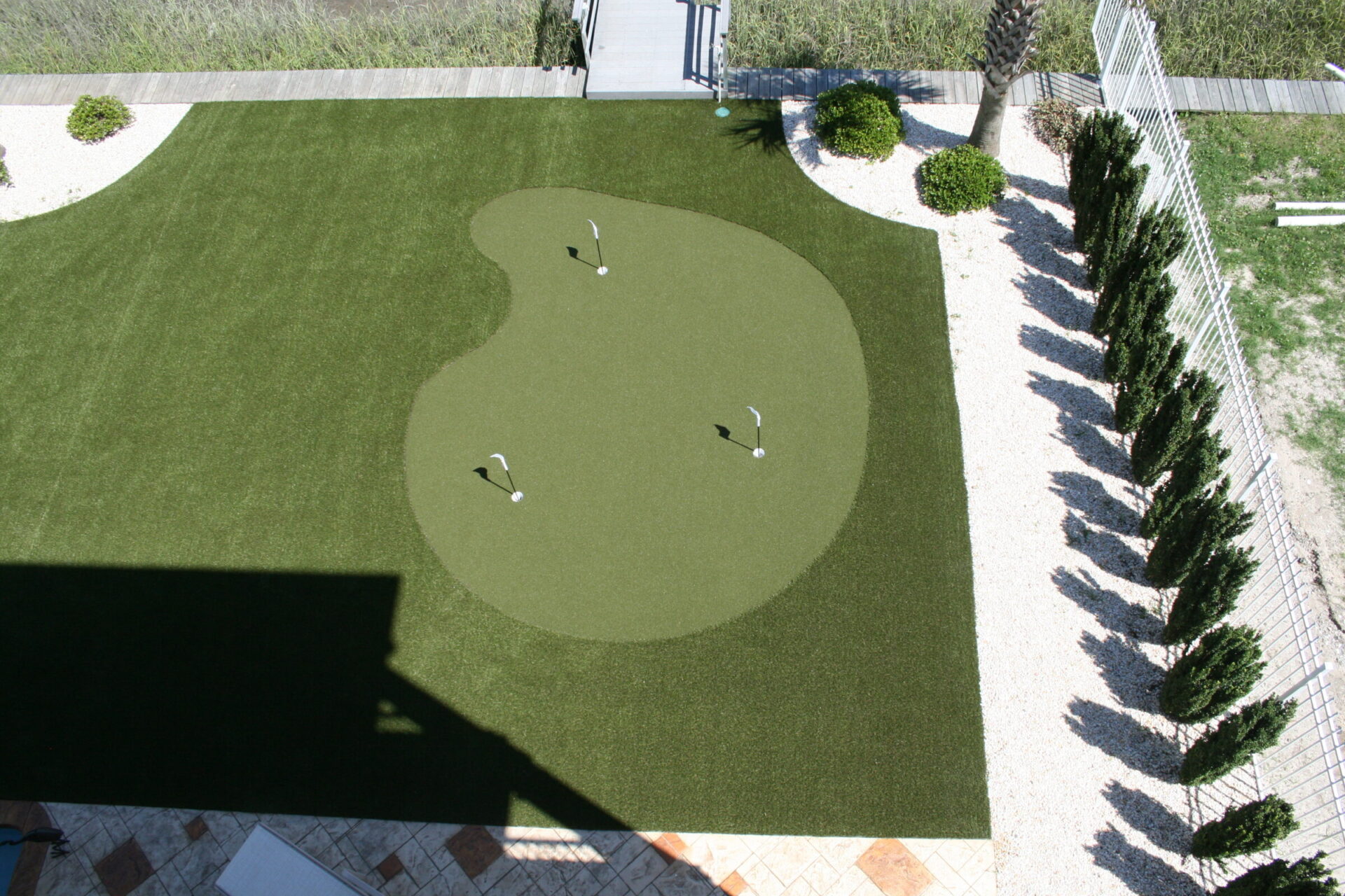 Aerial view of a private artificial turf putting green with three golf clubs and balls. Surrounded by a fence, trees, and landscaped areas.
