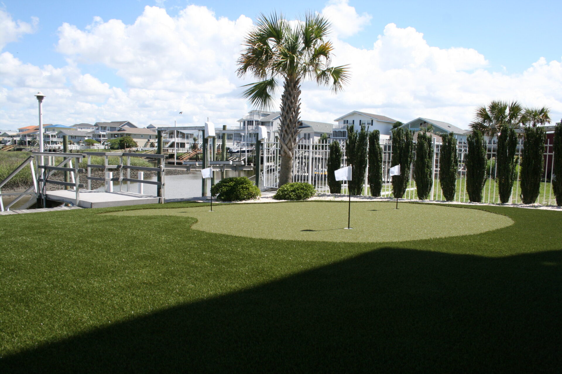 This is a well-manicured artificial putting green surrounded by a white fence and tall, thin trees, with a palm tree and residential area nearby.