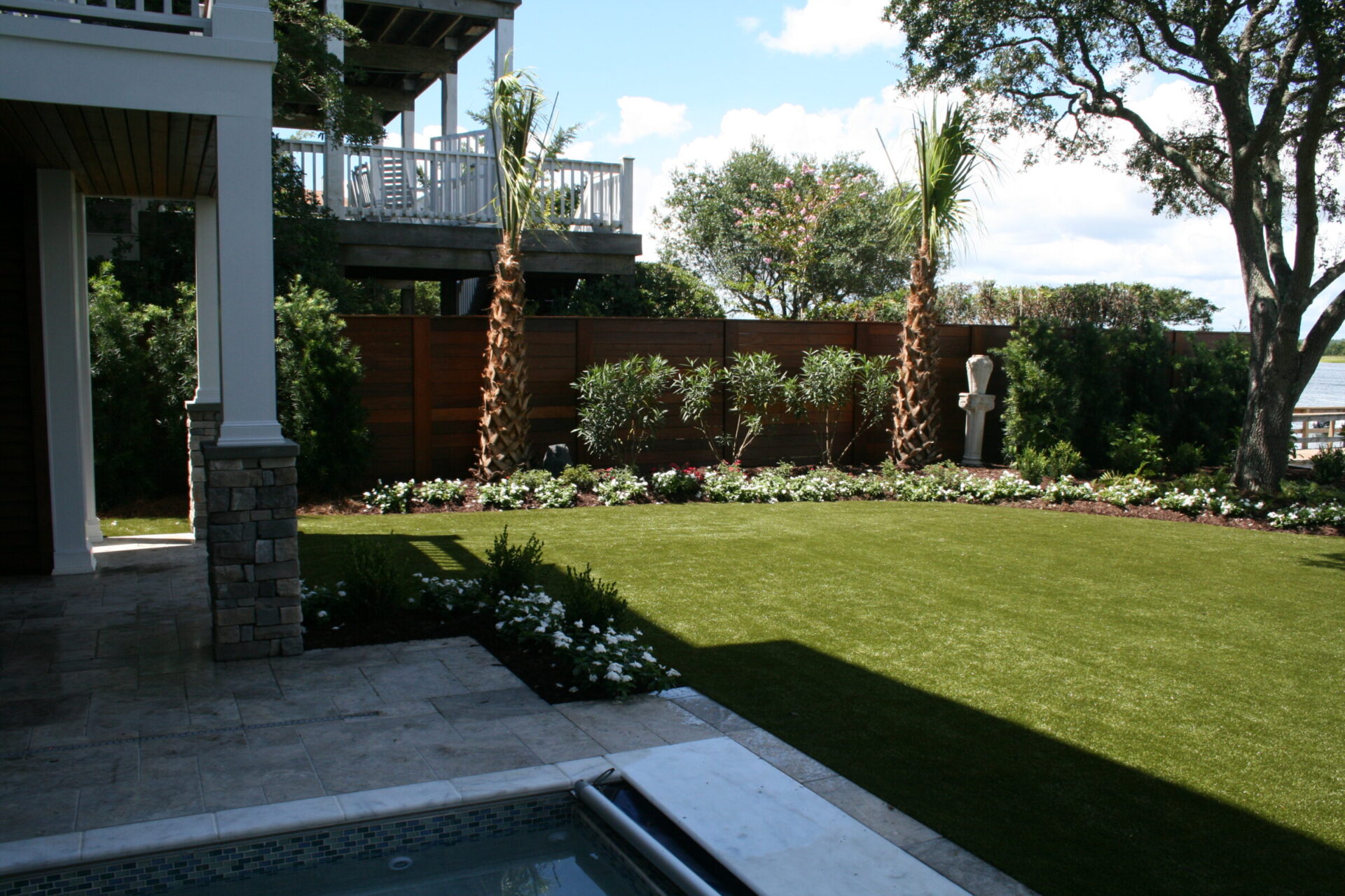 This image shows a beautifully landscaped backyard with a pool, neat lawn, tall palm trees, flowering plants, and a view of a water body.