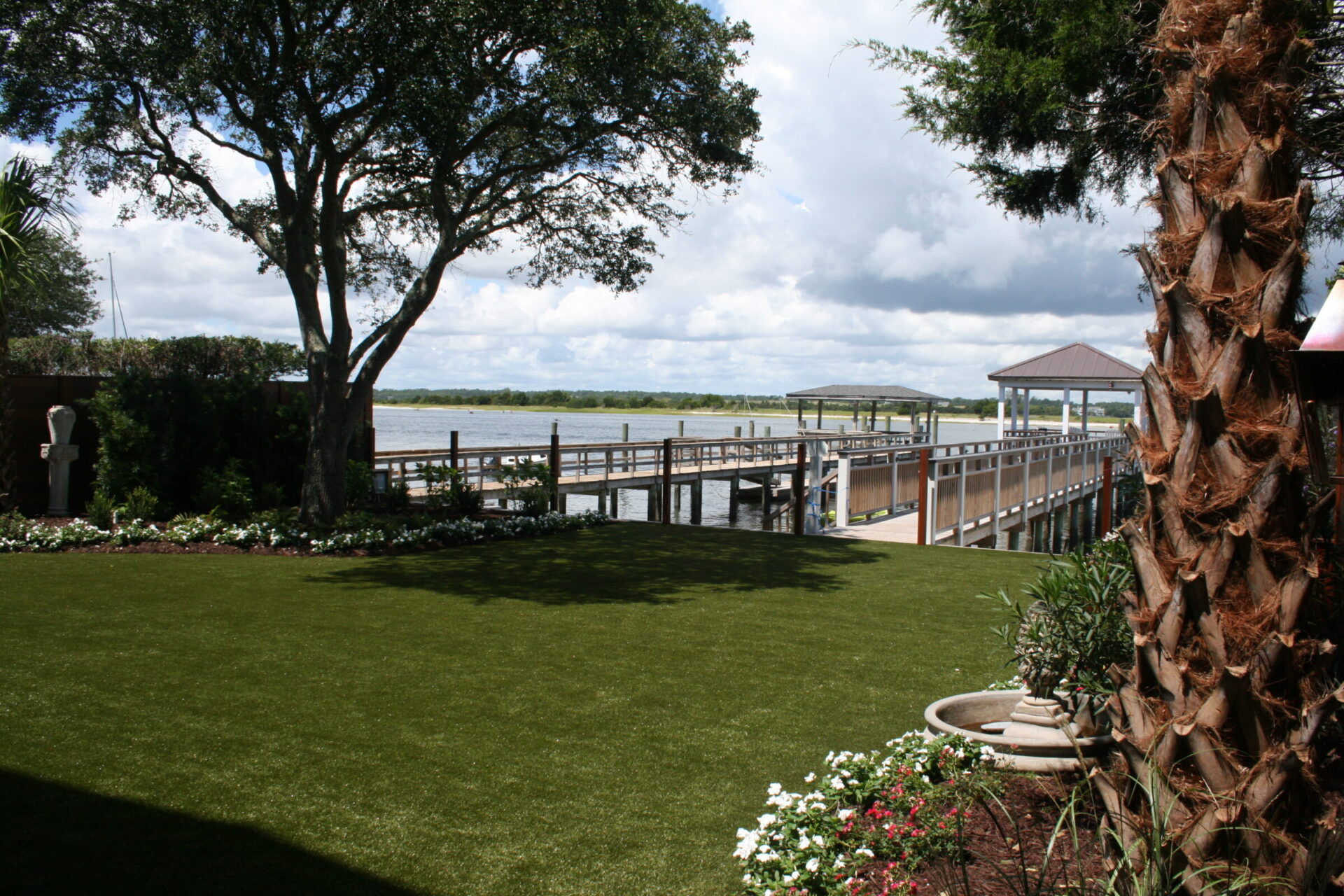 This image shows a lush garden with artificial turf, a large tree, and a wooden dock extending into a tranquil water body, with a cloudy blue sky above.
