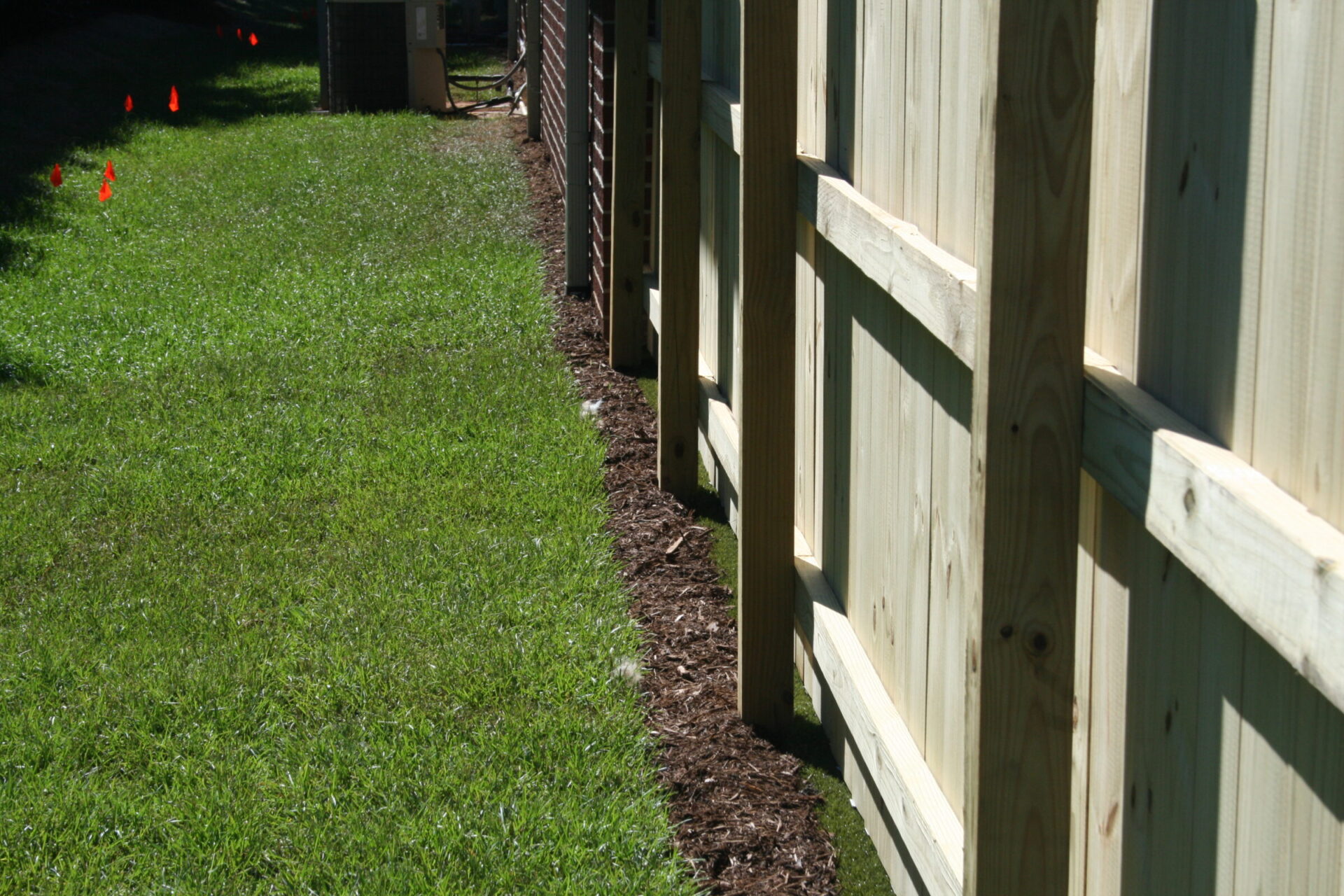 This image shows a neatly manicured grass lawn beside a wooden plank fence, with a line of mulch along the fence's base, in sunny daylight.