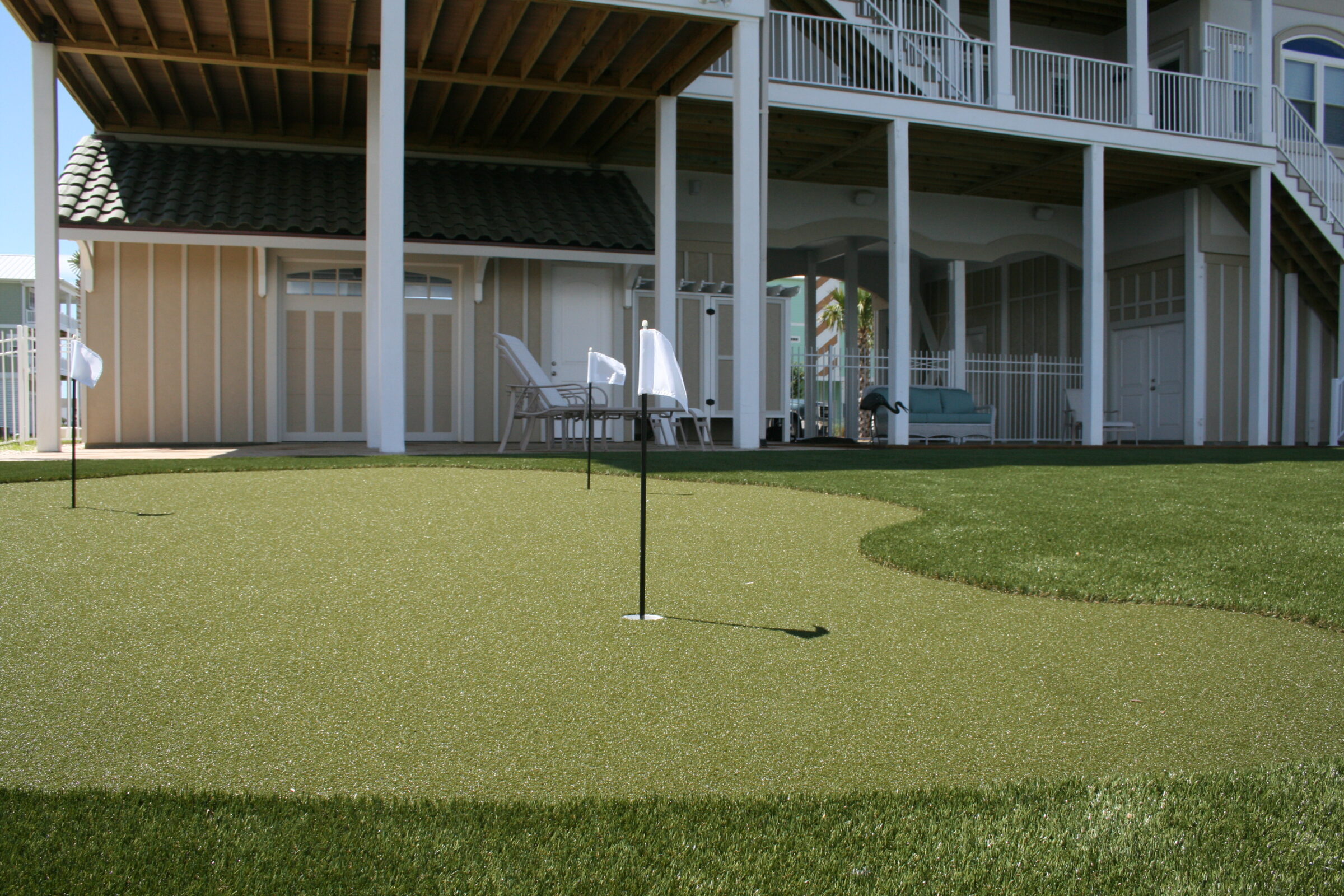 An artificial grass putting green with a golf hole and flag in front of a two-story house with a balcony and patio furniture. Clear blue sky overhead.