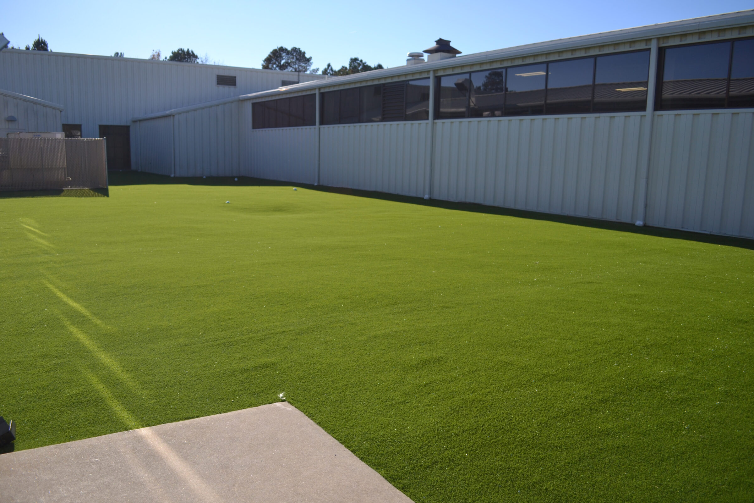 An outdoor area with artificial green turf, bordered by a metal building with large windows, and a clear sky above. The area appears unused.