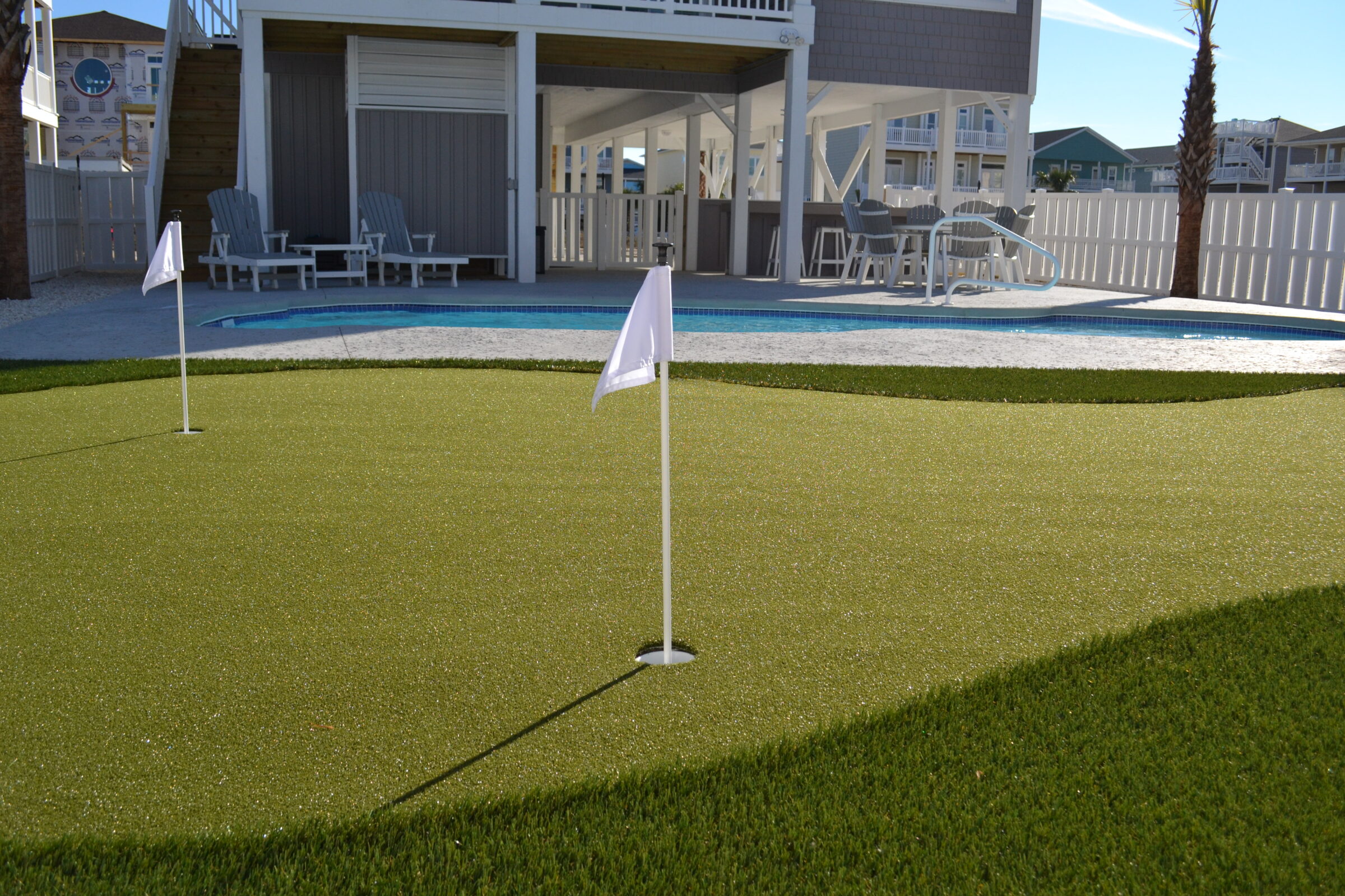 A backyard miniature golf green with two holes and flags, adjacent to a swimming pool and a house with outdoor furniture under a covered patio.