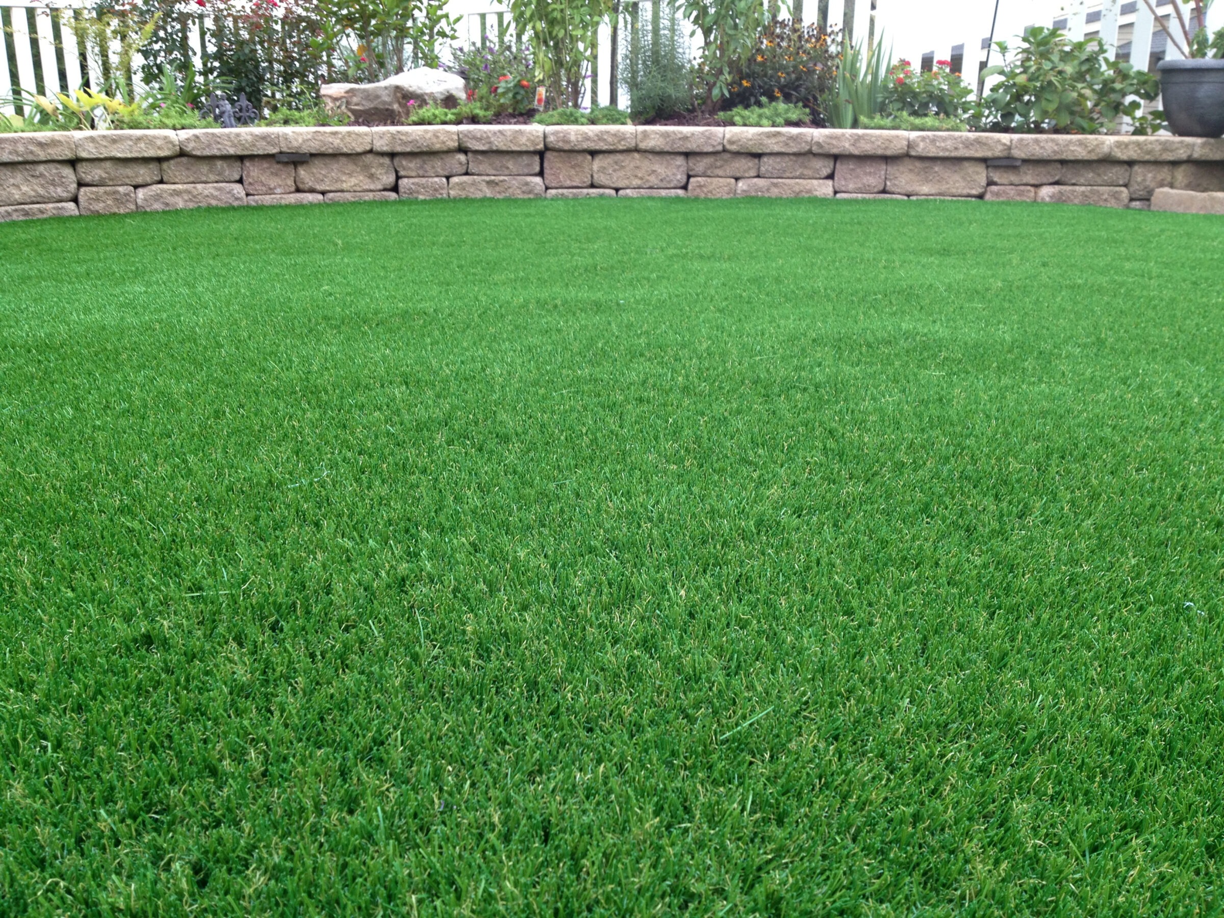A lush green artificial turf lawn is bordered by a curved stone retaining wall, with plants and a black pot visible in the background.