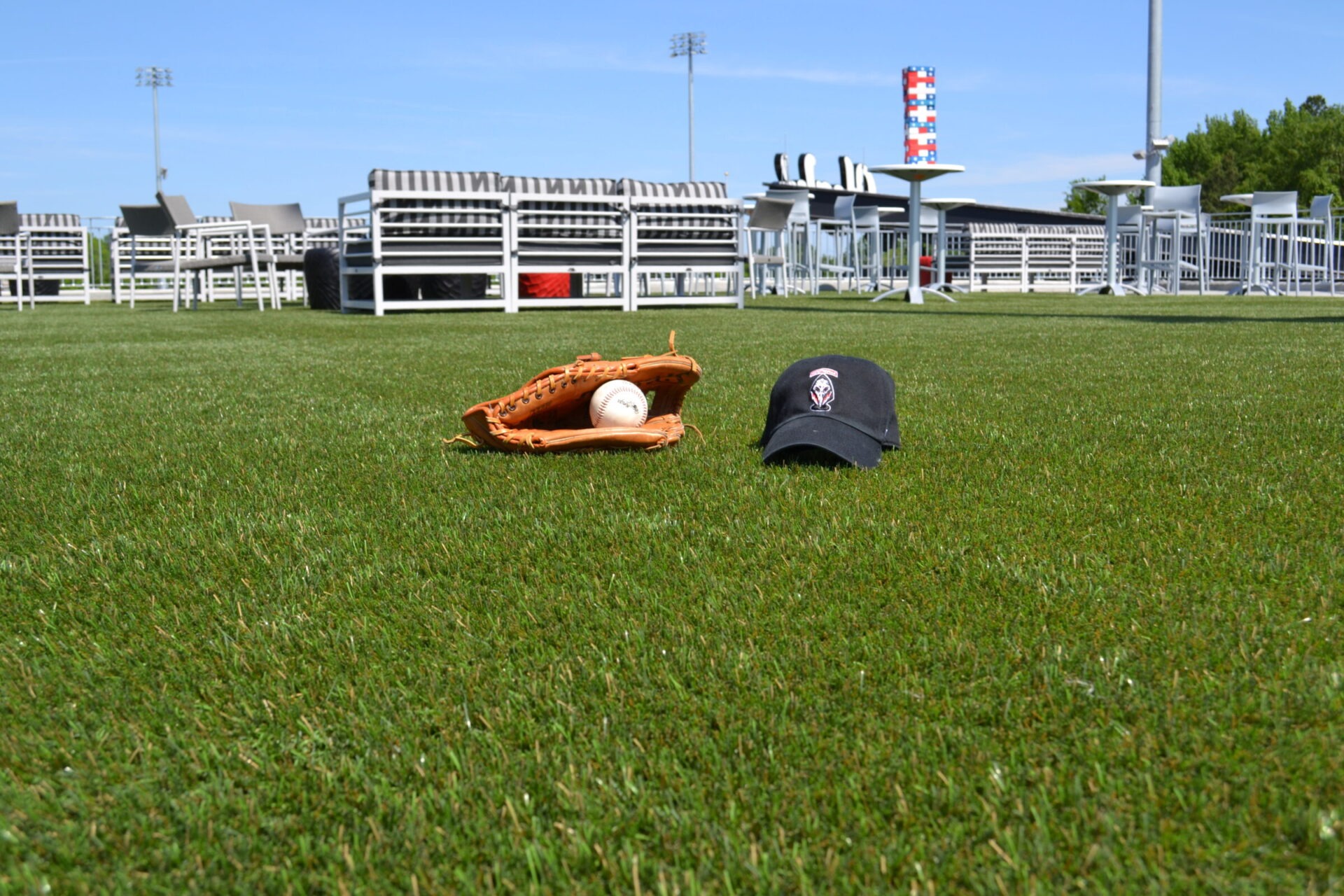 A baseball glove with a ball inside it and a black cap are on a lush green field, with empty bleachers and clear skies in the background.