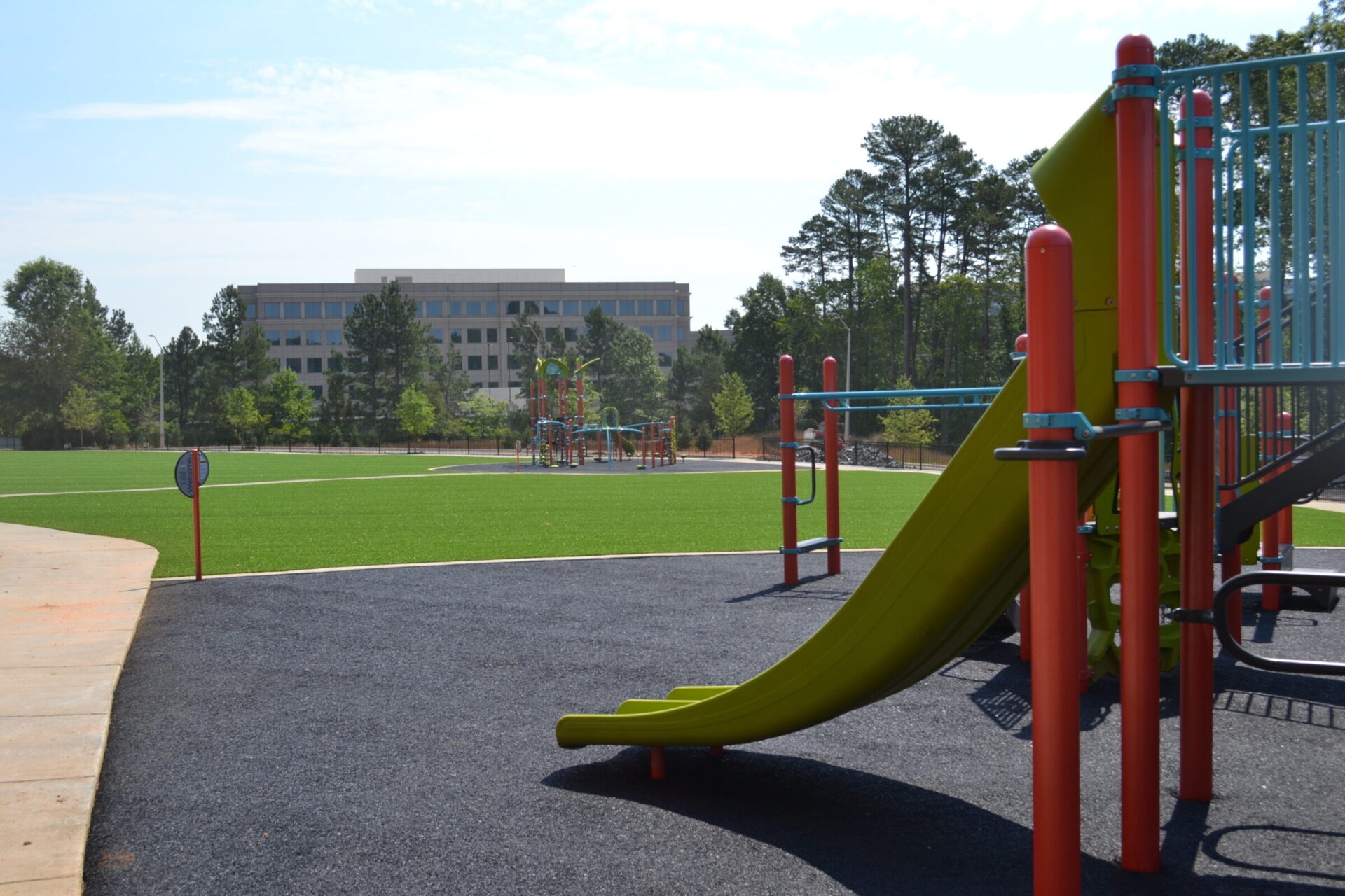 A colorful playground with green synthetic turf features a yellow slide, climbing structures, and outdoor fitness equipment, with trees and a building in the background.