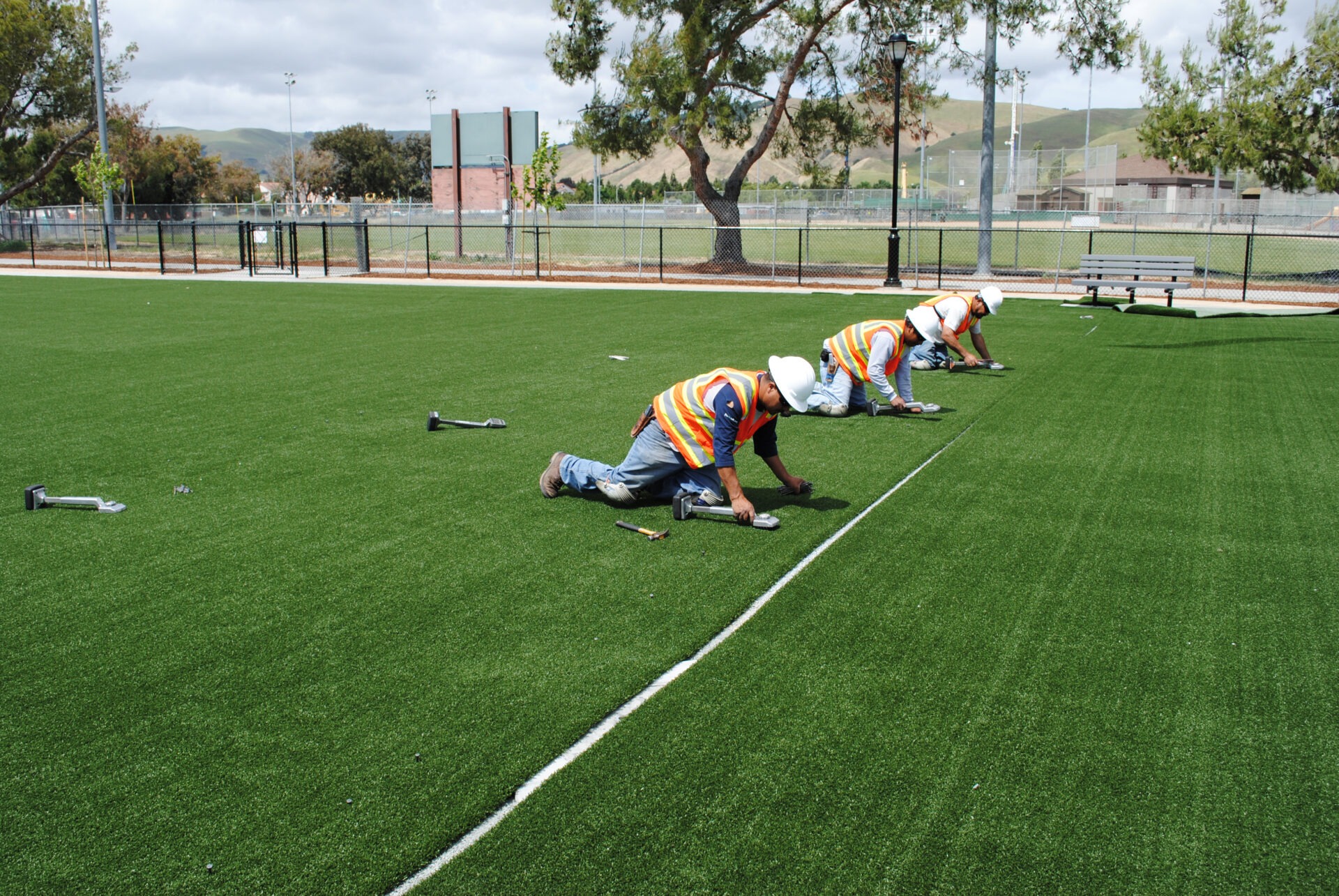 Three people in reflective vests and helmets are installing artificial turf on a sports field, with tools and a fence visible in the background.
