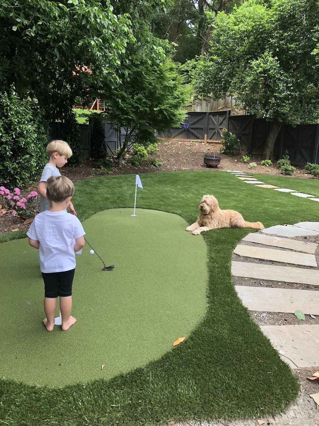 Two children and a dog are in a backyard with a small putting green. There's lush greenery, a fire pit, and stepping stones in the grass.