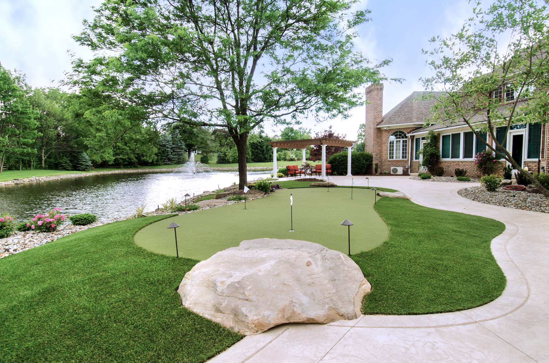 The photo shows a golf course putting green made of artificial turf, in front of a pond and fountain, near the entrance to a clubhouse.
