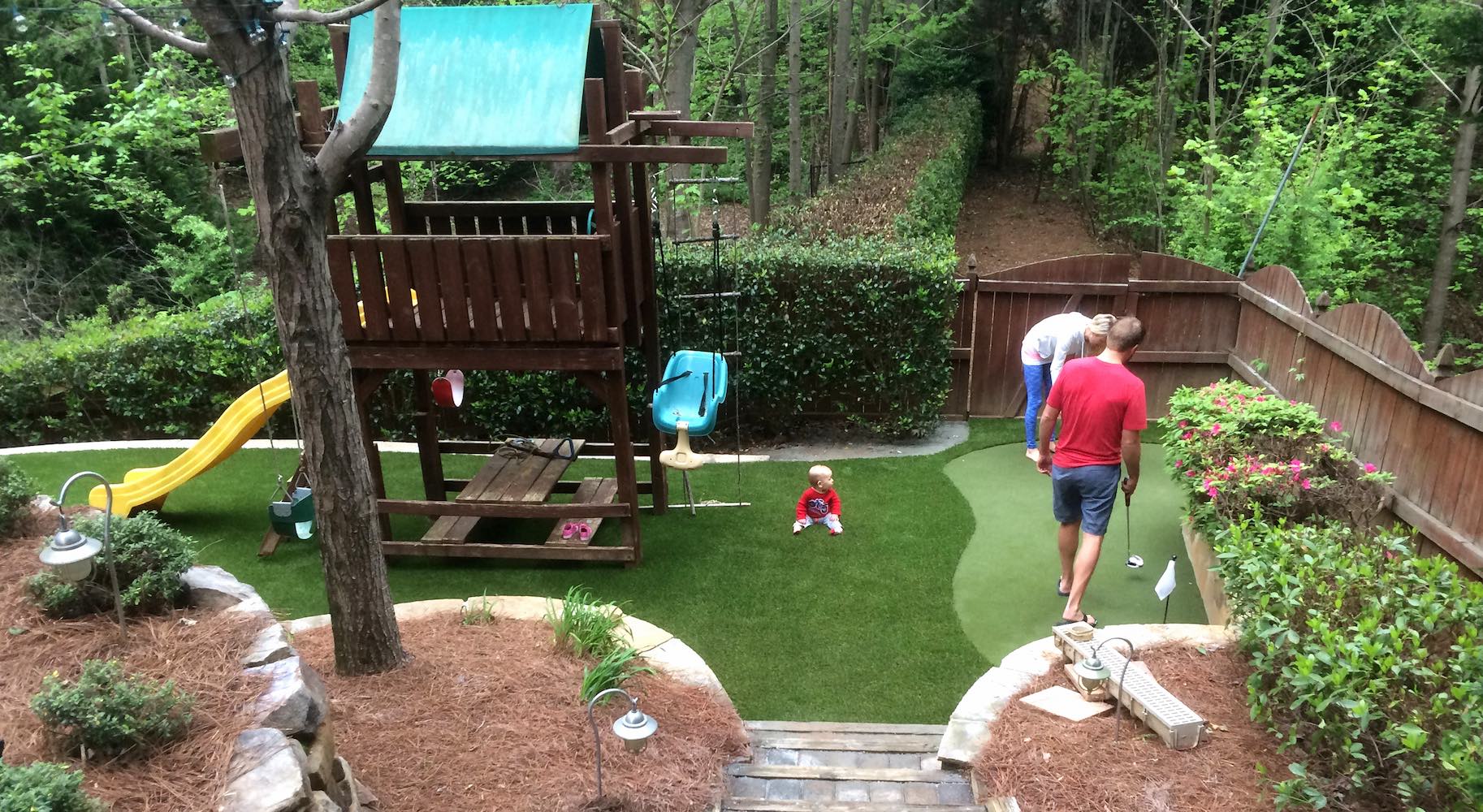 A person is playing miniature golf in a backyard with synthetic turf, landscaping, a swing set, and slide. Another person is watching a child nearby.