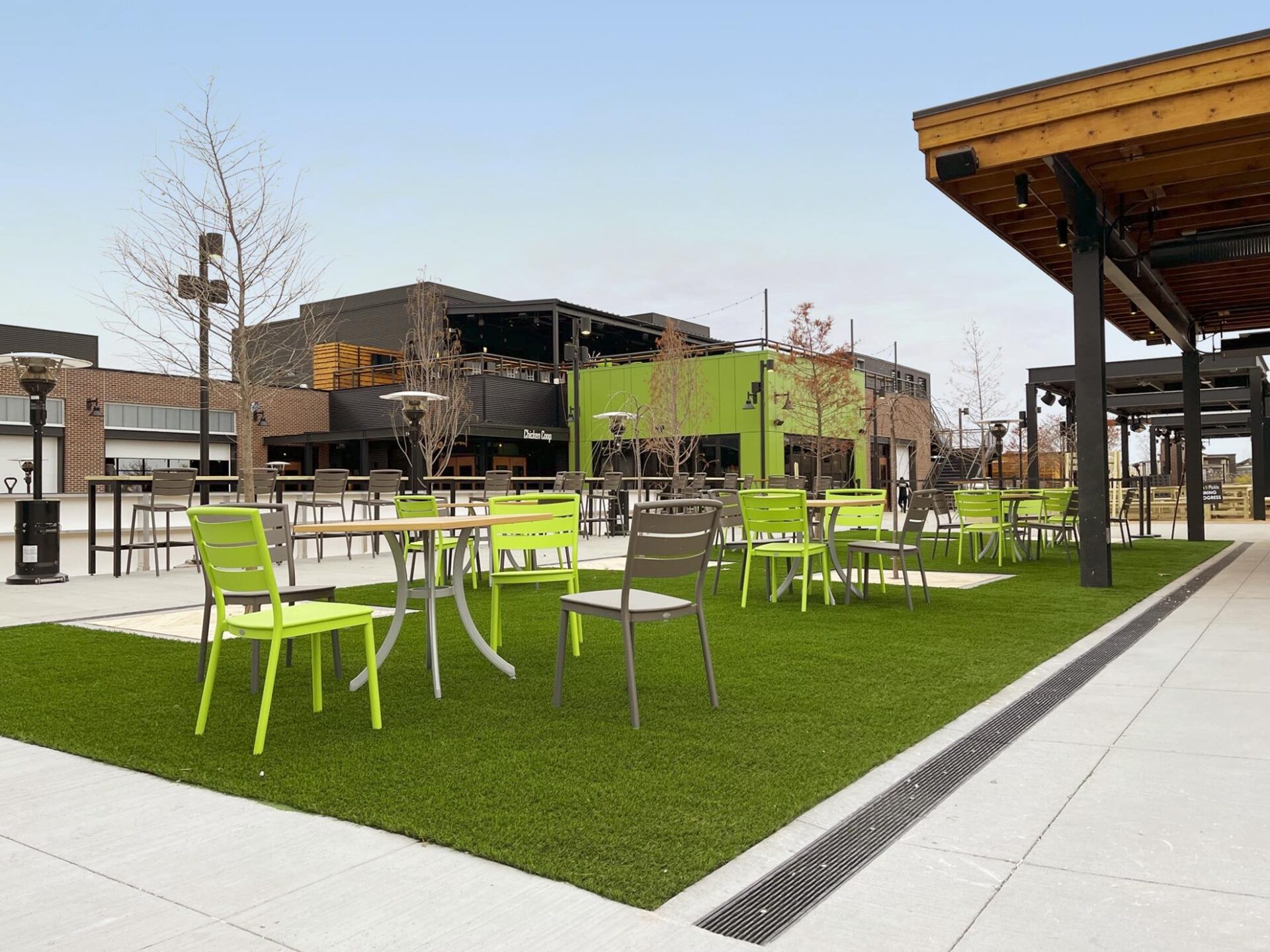 Outdoor dining area with vibrant green chairs and tables on synthetic grass, modern buildings in the background, and bare trees under a clear sky.