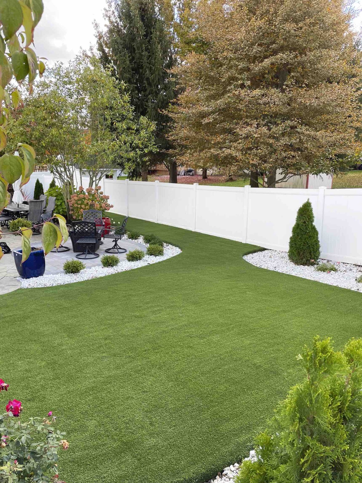 This image displays a neatly manicured garden with vibrant green artificial turf, surrounded by a white fence, various plants, trees, and garden furniture.