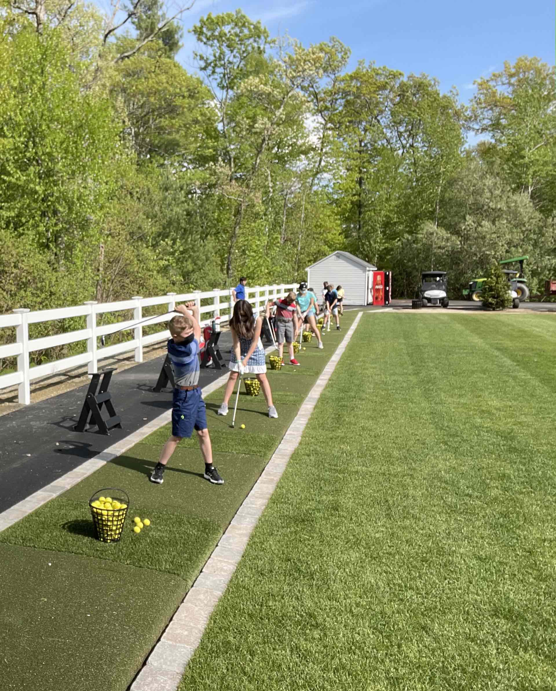 People, including a child, are practicing golf on a driving range with artificial turf mats, trees in the background, and golf carts to the side.