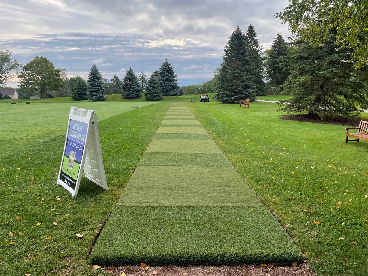 A golf driving range with artificial turf mats aligned in a row, a sign advertising golf lessons, trees, grass, and a bench under an overcast sky.