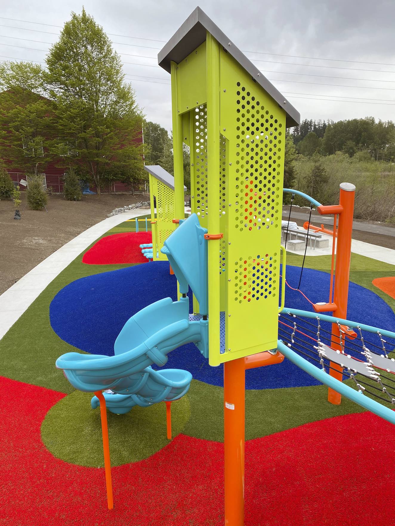 Brightly colored playground equipment on vibrantly hued safety surfacing, featuring a blue slide, climbing structures, and greenery in the background.