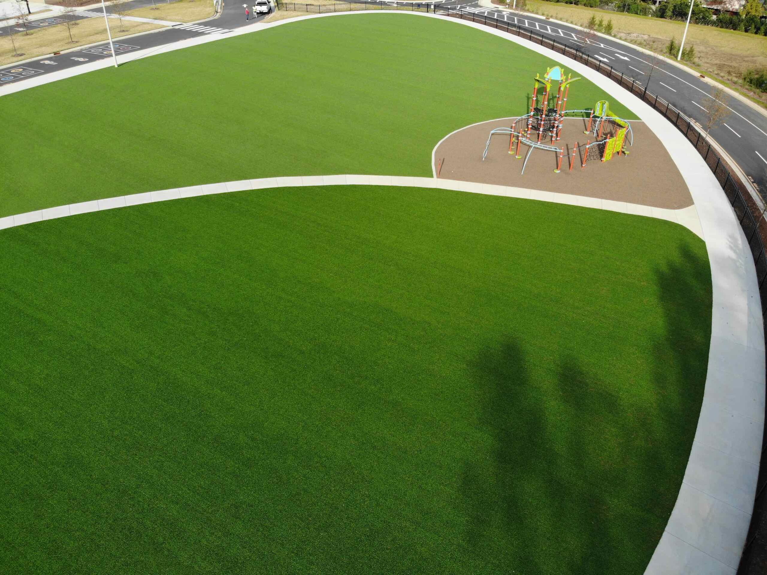 An aerial view of a playground with colorful equipment on brown safety surfacing, bordered by a curved white path against a green artificial turf.