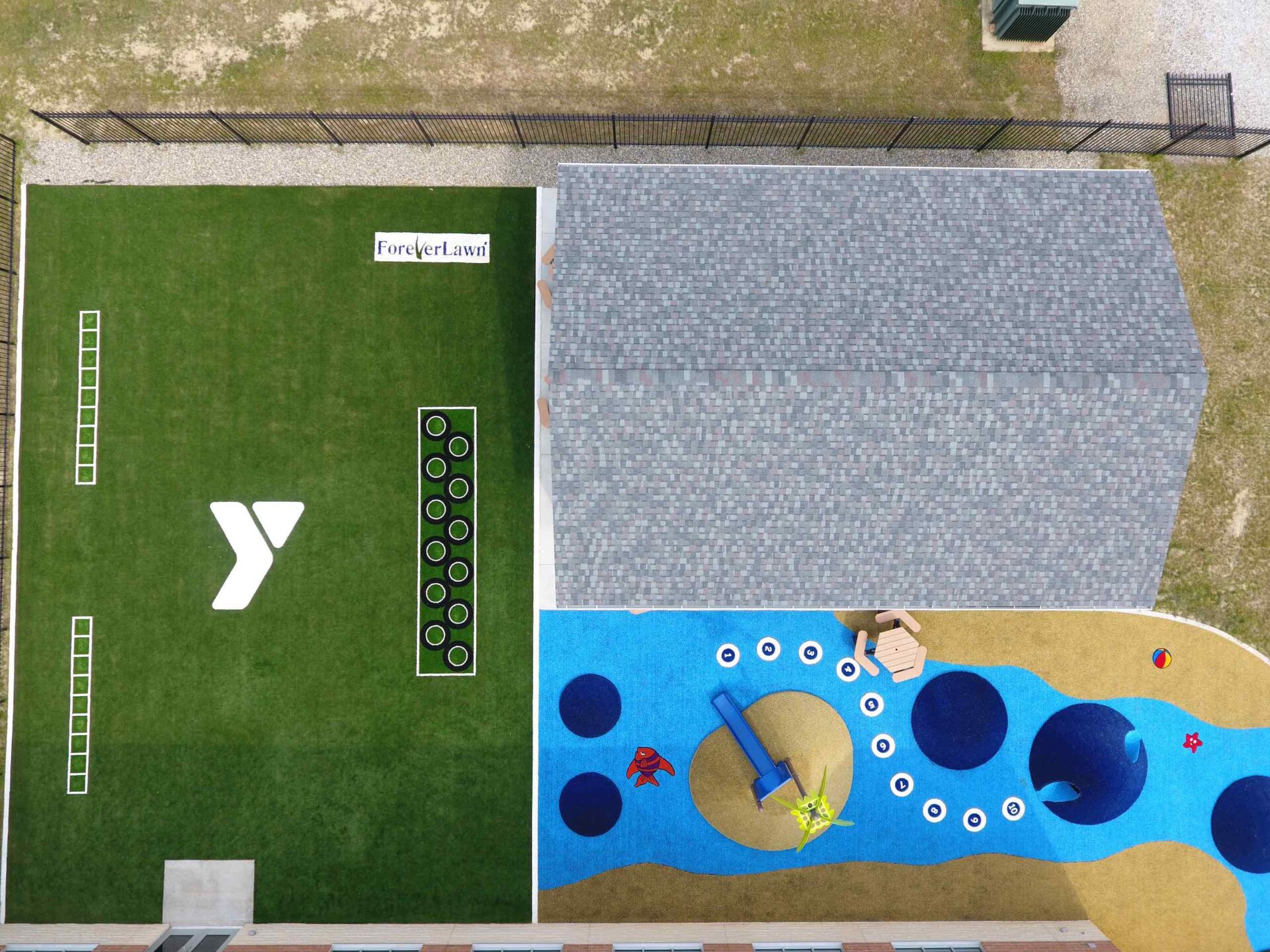 An aerial view of a playground with artificial grass, colorful play areas, a blue splash pad, and a shaded bench next to a building.