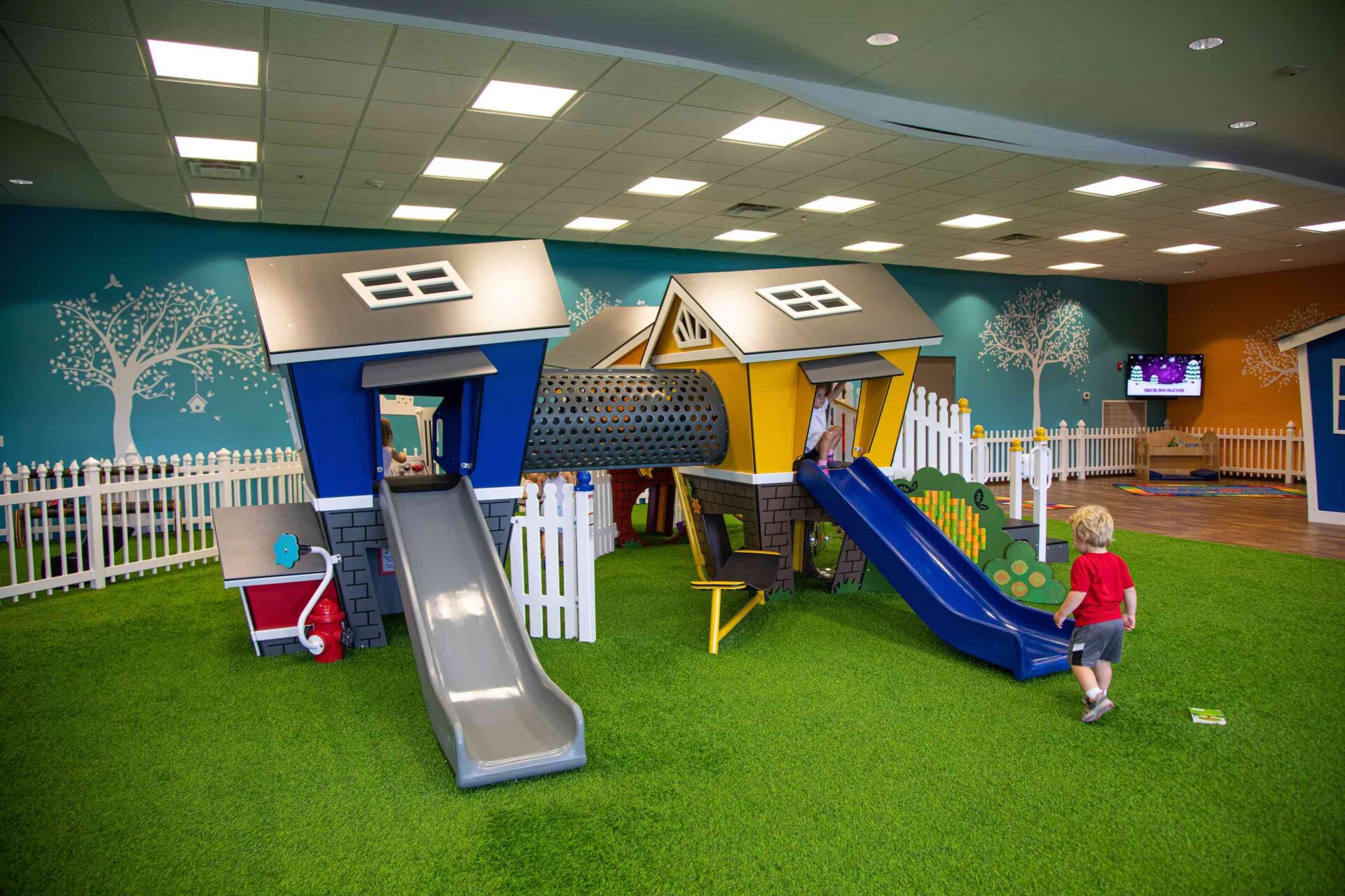 A colorful indoor playground with a green floor resembling grass, featuring slides, a playhouse, and a child about to climb a slide.