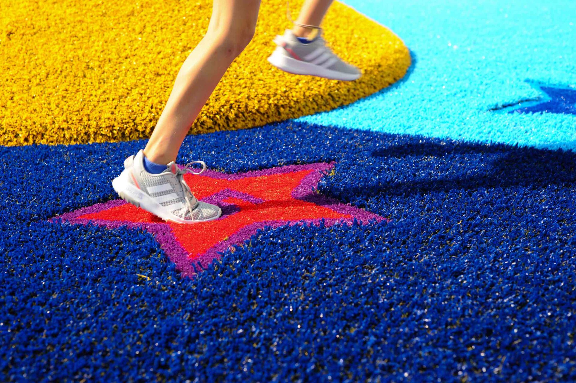 The image shows the legs of a person walking across a textured surface with bright primary colors and a red star pattern, likely at an outdoor event.