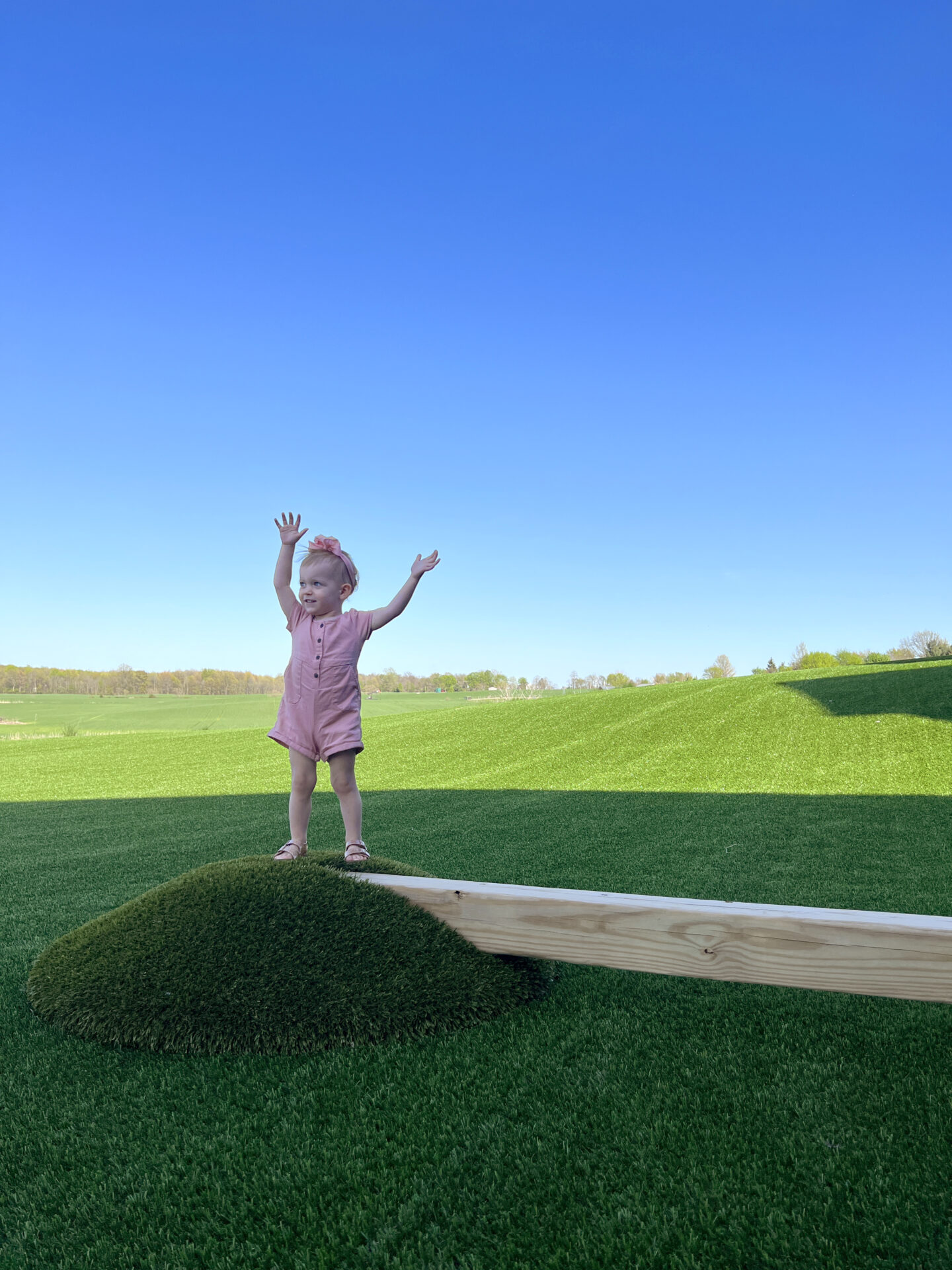 A child stands on an artificial turf hill against a blue sky, arms raised high, wearing a pink dress and sandals, expressing joy.