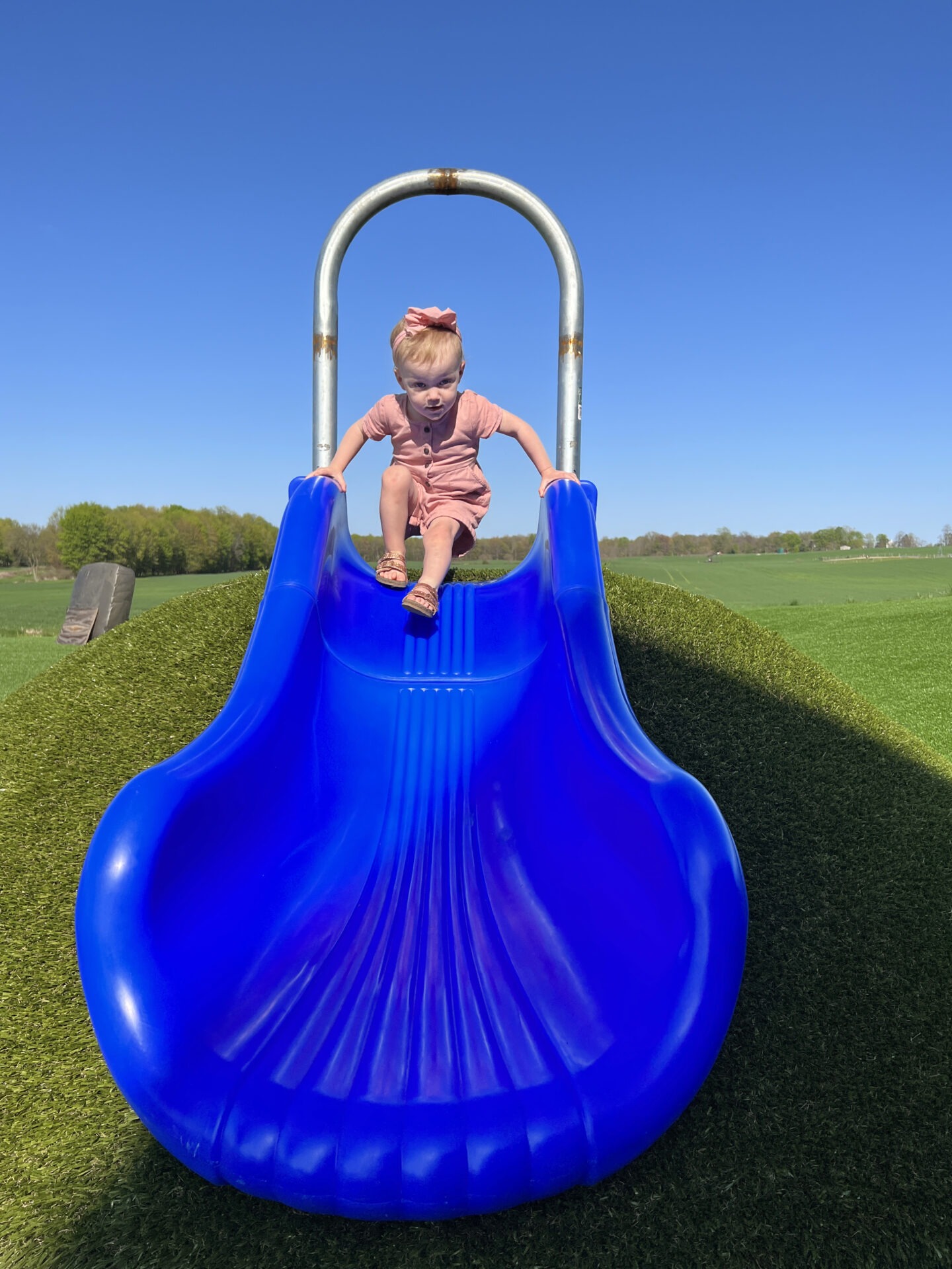 A young child poised at the top of a vibrant blue playground slide, with clear blue skies and open green fields in the background.