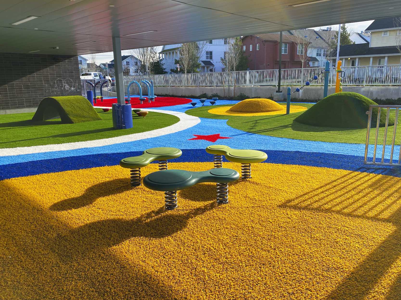 A colorful playground with artificial grass, vibrant safety surfacing, a four-seat spring teeter-totter, and play structures including slides and climbing features.