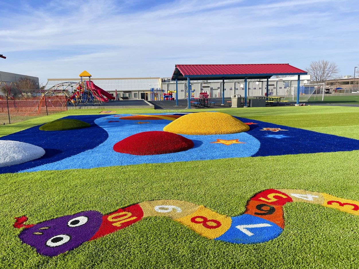 A colorful playground with artificial turf, vibrant play equipment, a counting caterpillar design on the ground, and a blue sky. No visible people.