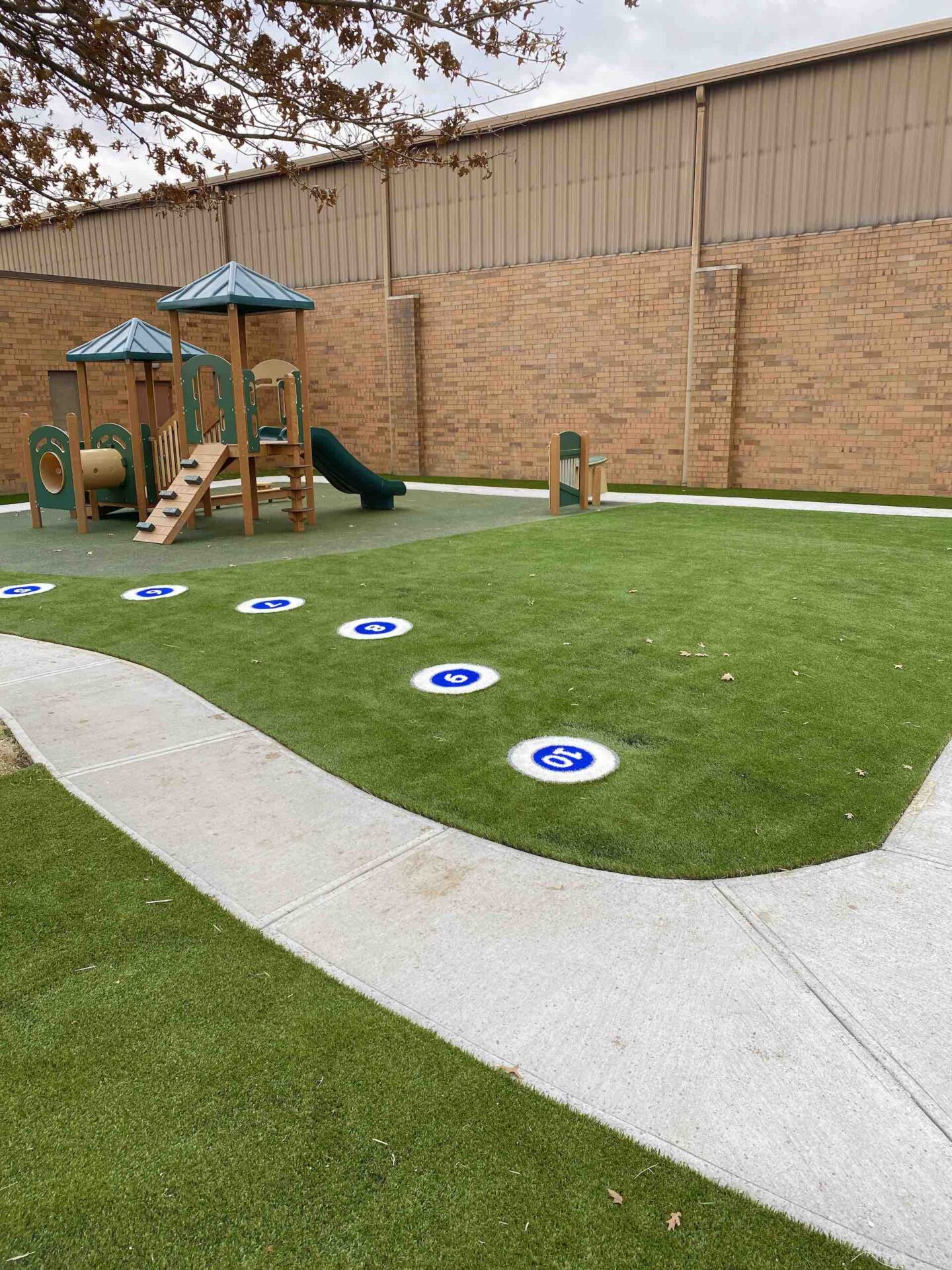 A playground with artificial grass features a slide structure, number-marked stepping stones, and a winding concrete path against a brick wall backdrop.