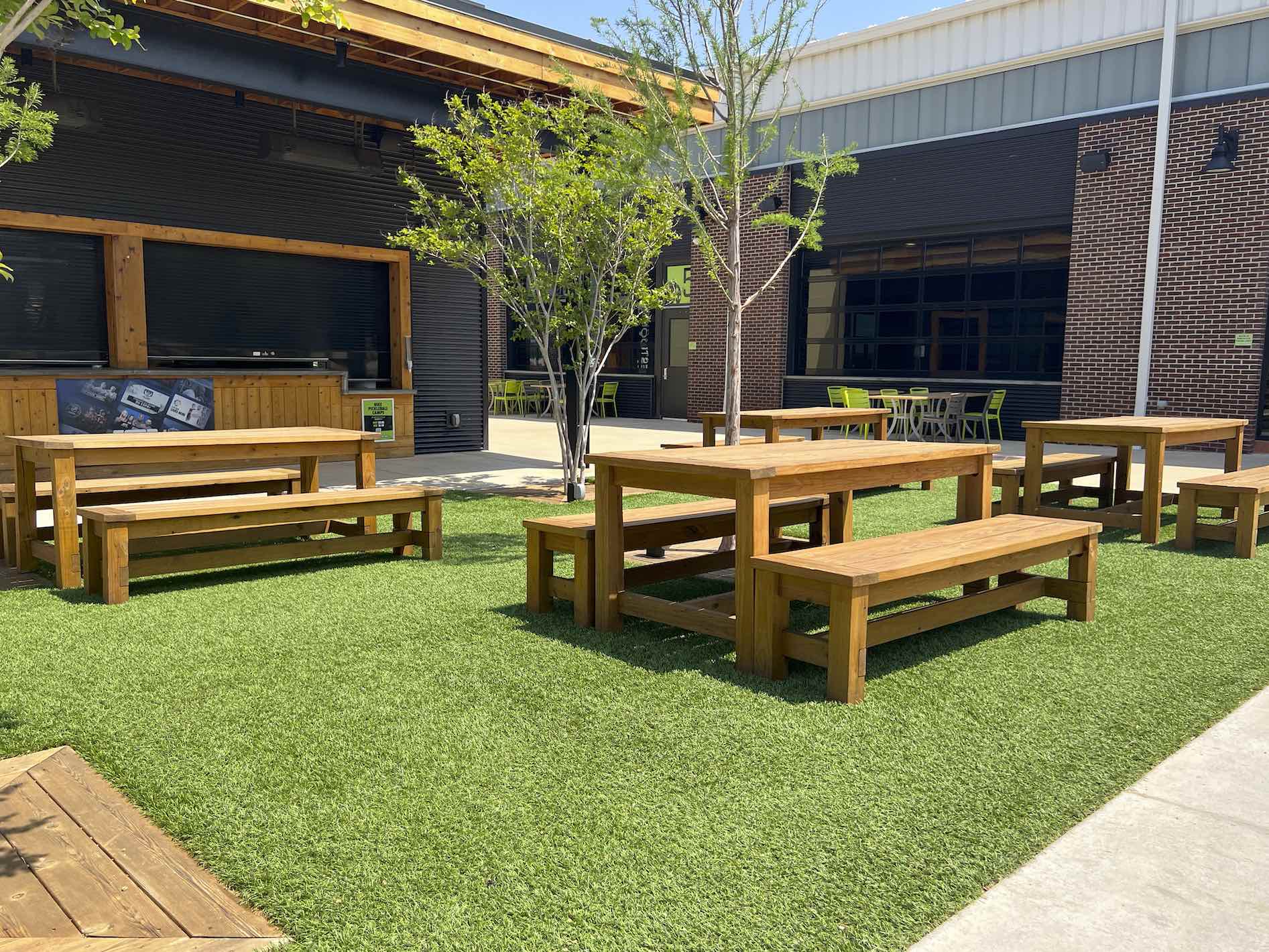 Outdoor patio with artificial grass, wooden tables and benches, a young tree, buildings with black and brick facades, and clear blue sky above.
