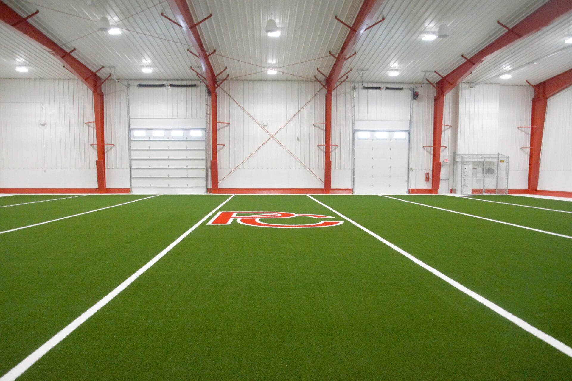 An indoor artificial turf field with white lines and a large logo at the center, within a large white building with red supports.