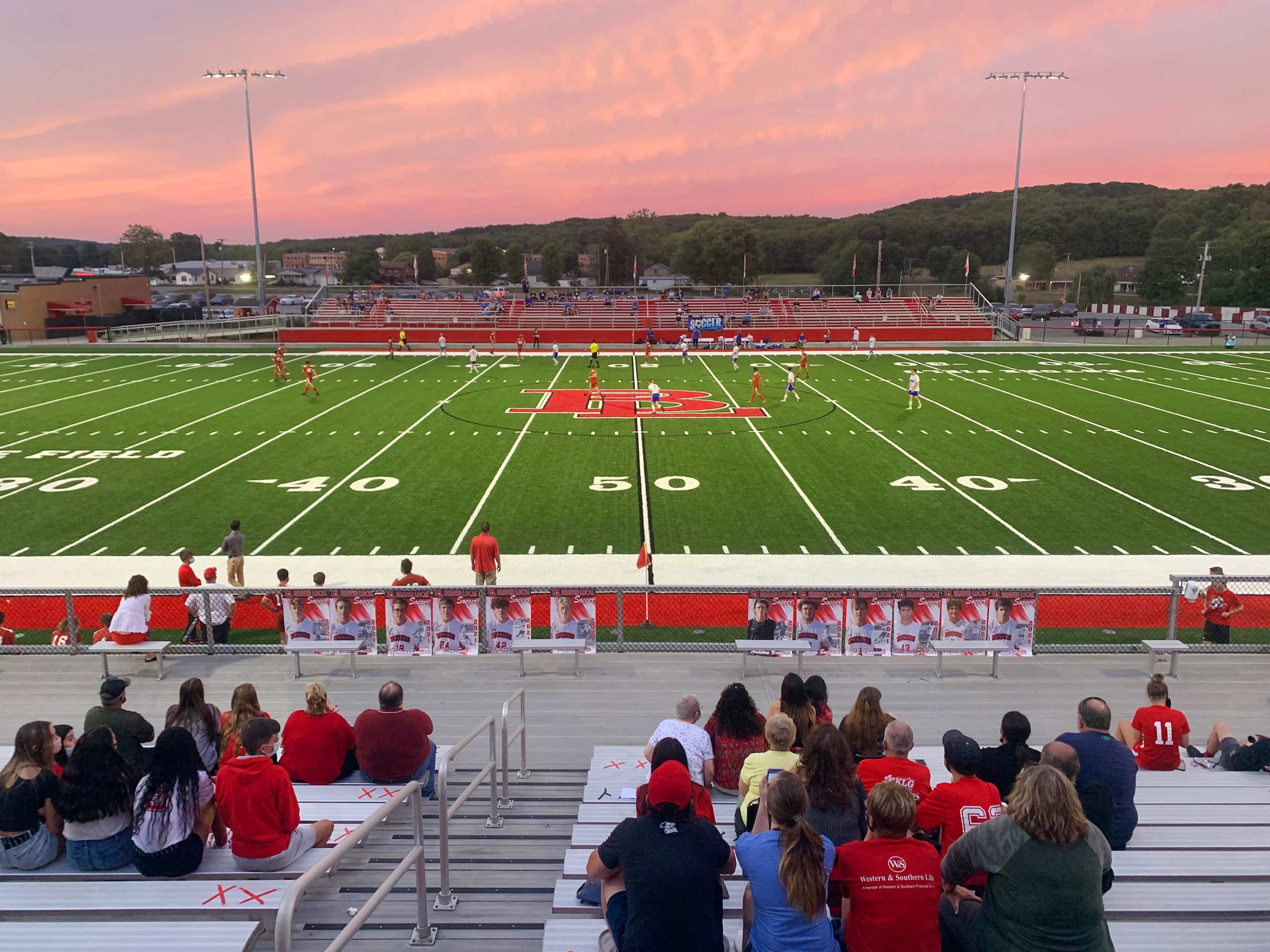 Spectators are seated in stadium bleachers watching a soccer match on a synthetic turf field during a vibrant sunset with a pink-hued sky overhead.