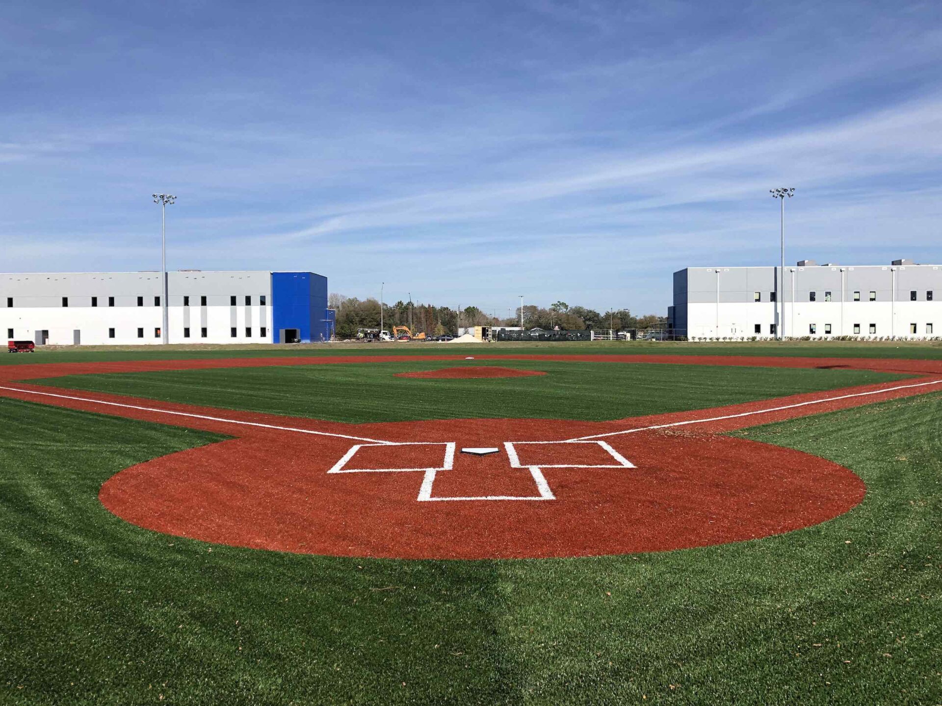 A pristine baseball field with red dirt infield, white chalk lines, and green outfield grass under a clear blue sky, flanked by large industrial buildings.