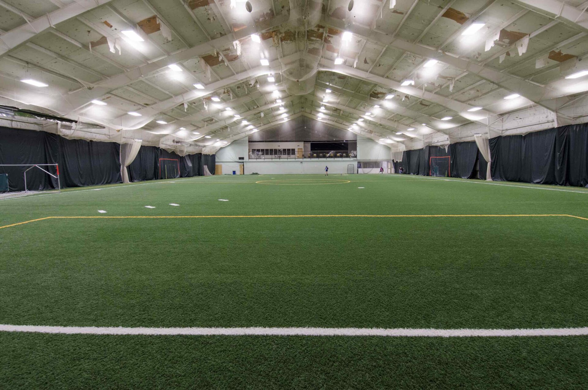 An indoor sports facility with green synthetic turf, white and yellow markings, a person running, and ceiling-mounted lights illuminating the space.