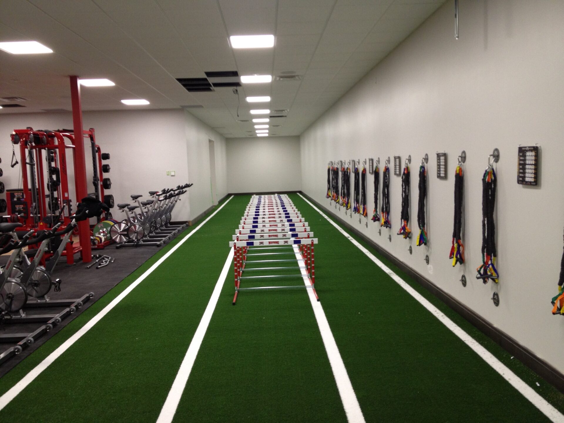 An indoor gym with a synthetic turf track, strength-training equipment, stationary bikes, resistance bands, and an agility ladder. The facility appears clean and organized.