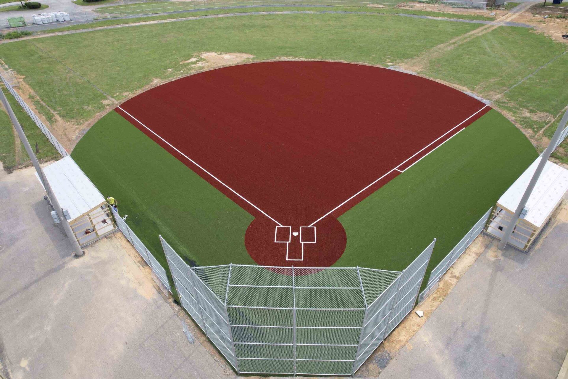 Aerial view of an empty baseball field with red and green artificial turf, white lines, and dugouts, surrounded by a chain link fence.