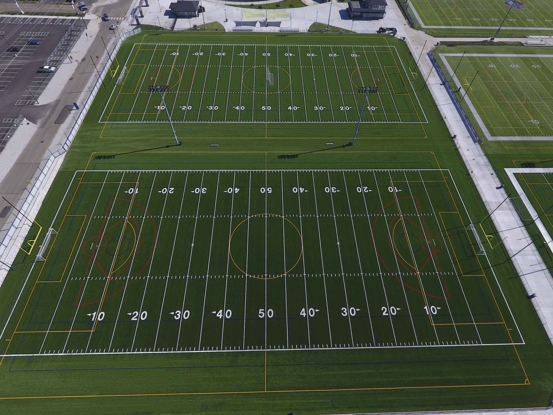 An aerial view of multiple sports fields with various line markings for football and soccer, surrounded by parking spaces and empty roads.